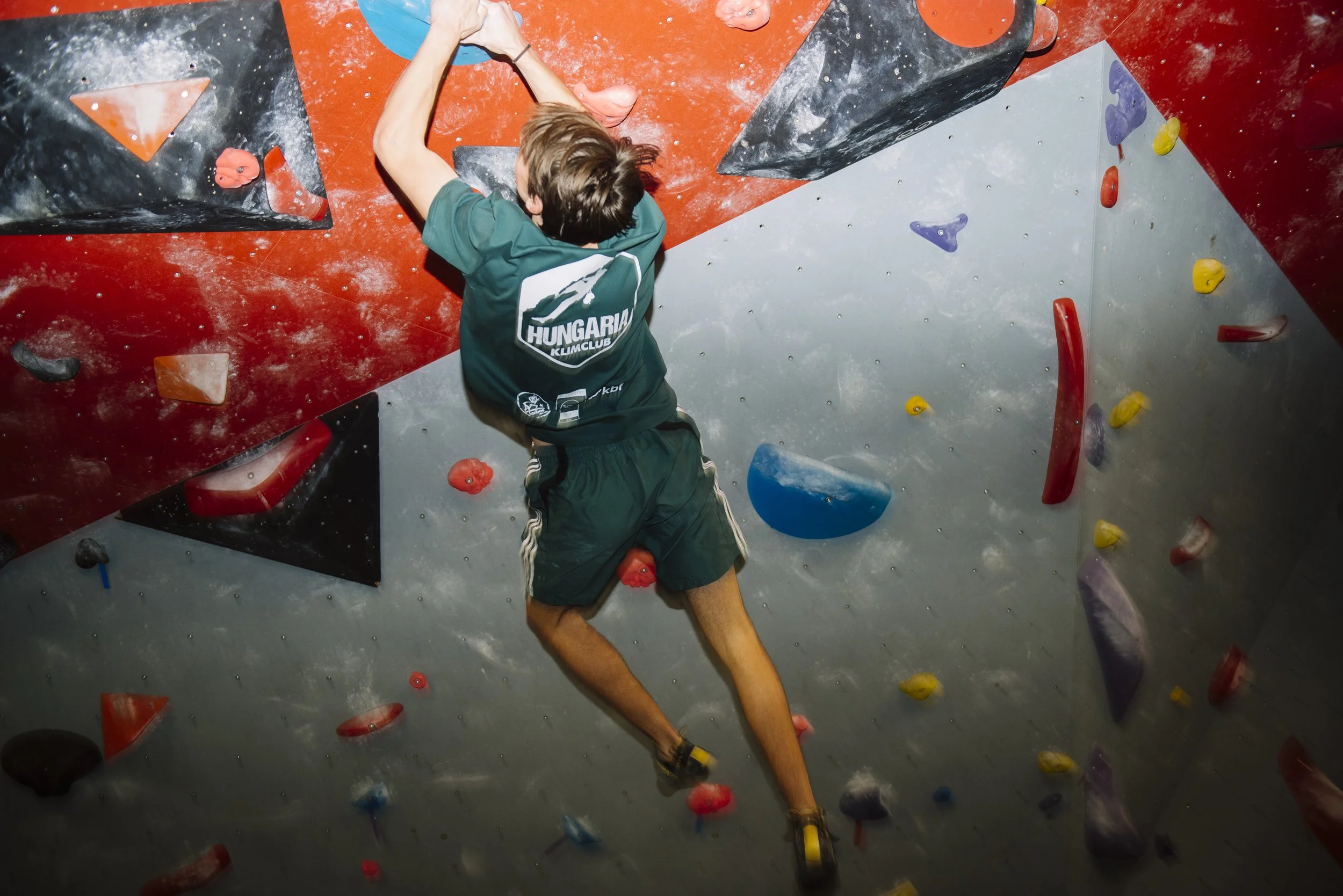 een jongen die supereenvoudig klimt in een indoorsporthal, gekleed in groene sportkleding, met kleurrijke klimwanden en -grepen.