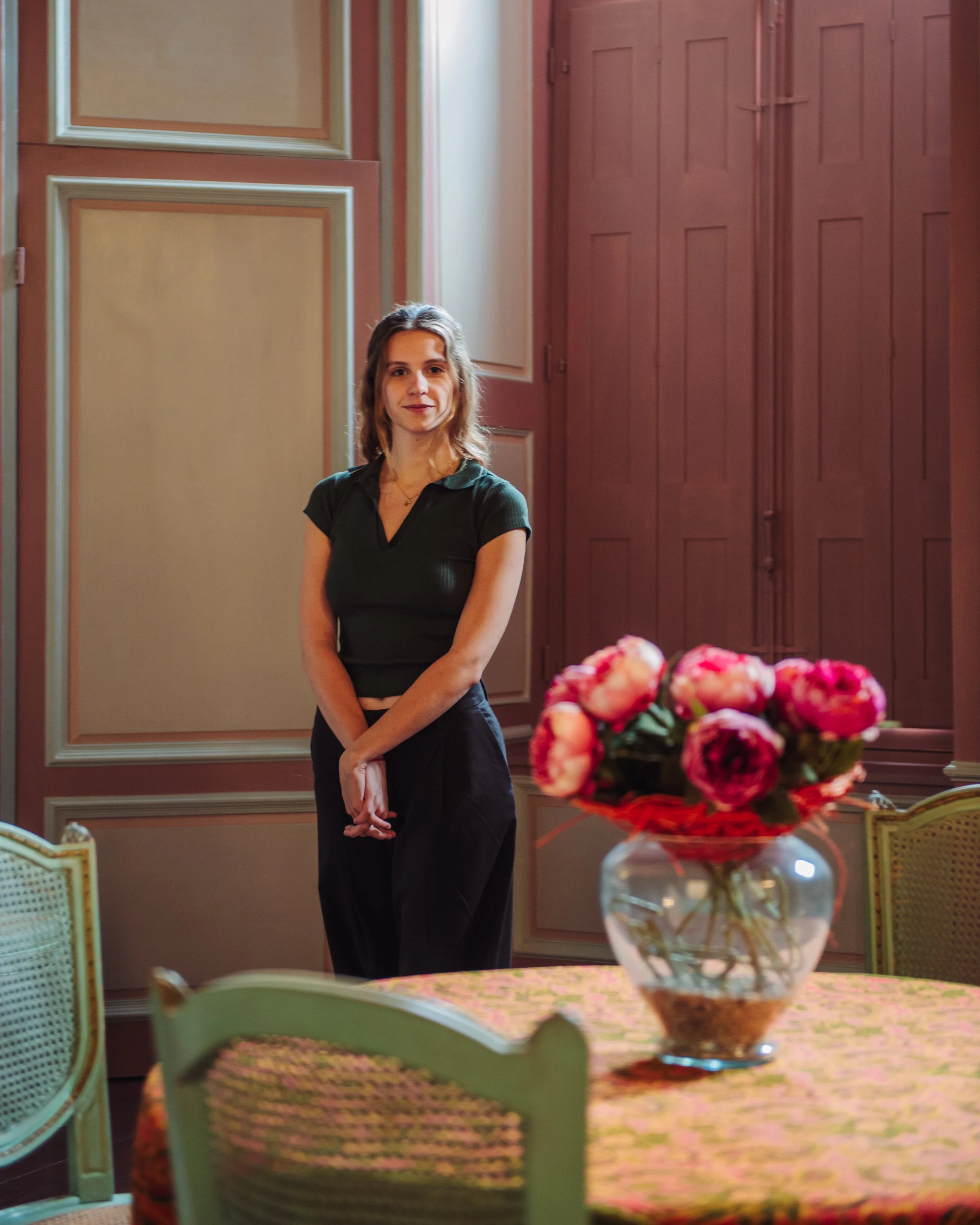 Een jonge vrouw met bruin haar in een zwart overhemd en zwarte broek staat in een kamer met roodbruine houten panelen en een tafel met een vaas met roze bloemen.