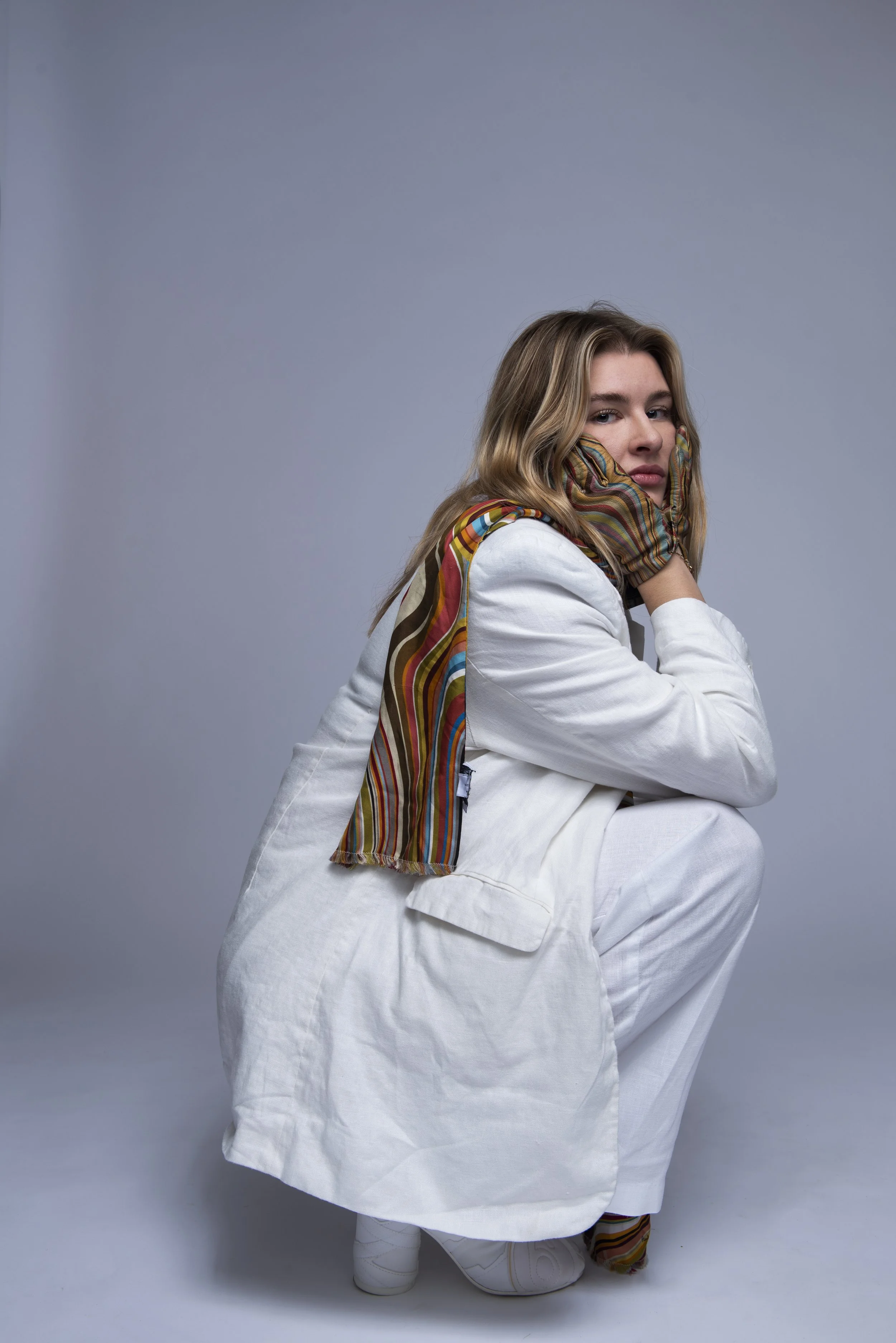 Een jonge vrouw in een witte jas en kleurrijke sjaal, zittend op de grond, met haar kin rustend op haar hand, tegen een grijze achtergrond.