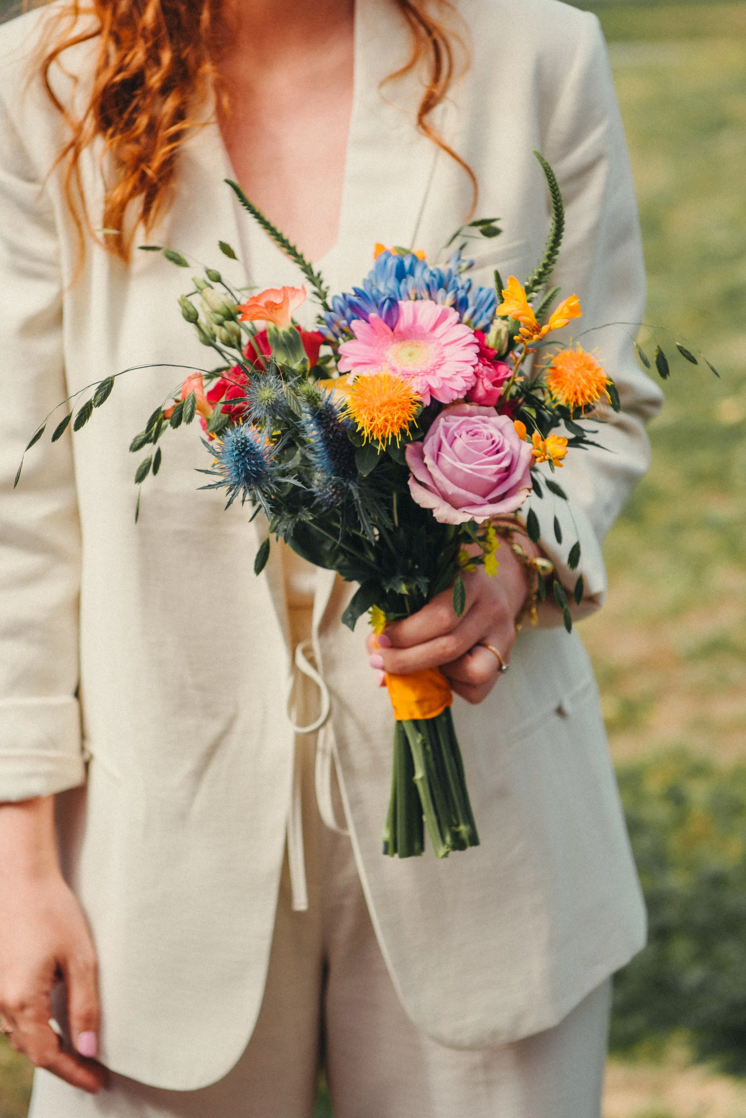 Een persoon houdt een kleurrijk boeket bloemen vast, bestaande uit rozen, asters en andere bloemen, met een oranje lint om de stelen. De persoon draagt een lichtkleurige blazer. De achtergrond is een groene, natuurlijke omgeving.