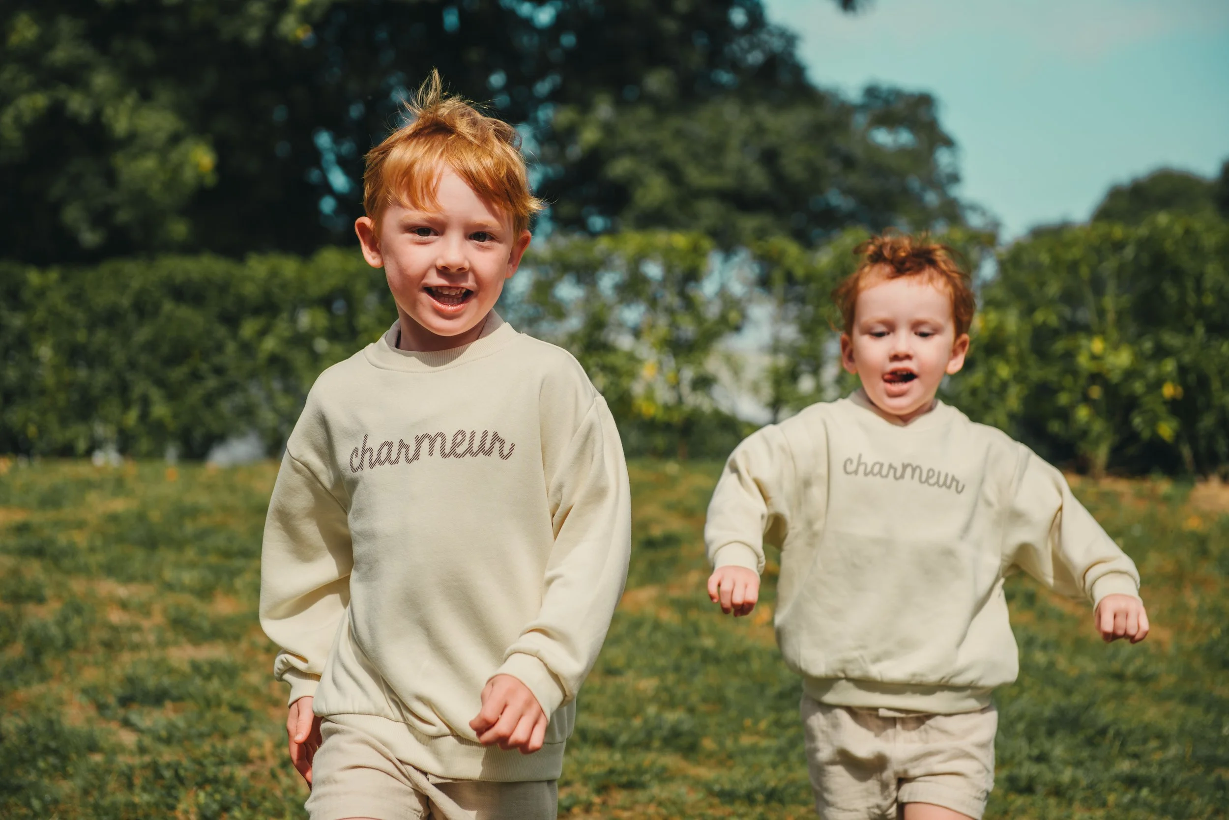Twee jonge kinderen rennen en spelen buiten in een groene tuin, gekleed in lichtkleurige sweatshirts met het woord 'charmeur' erop.
