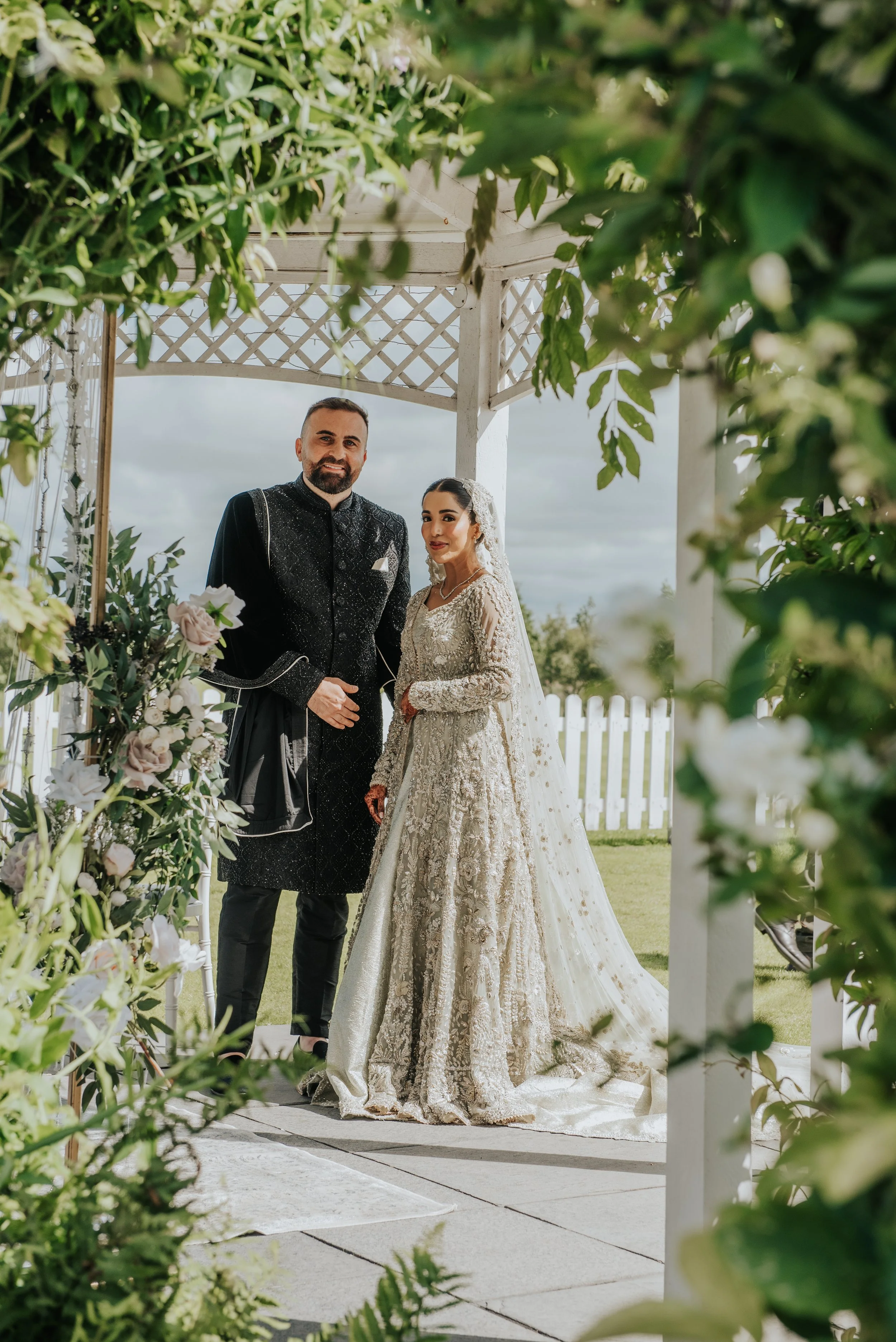 Bruid en bruidegom poseren samen onder een witte boog met bloemen tijdens een bruiloft buiten.