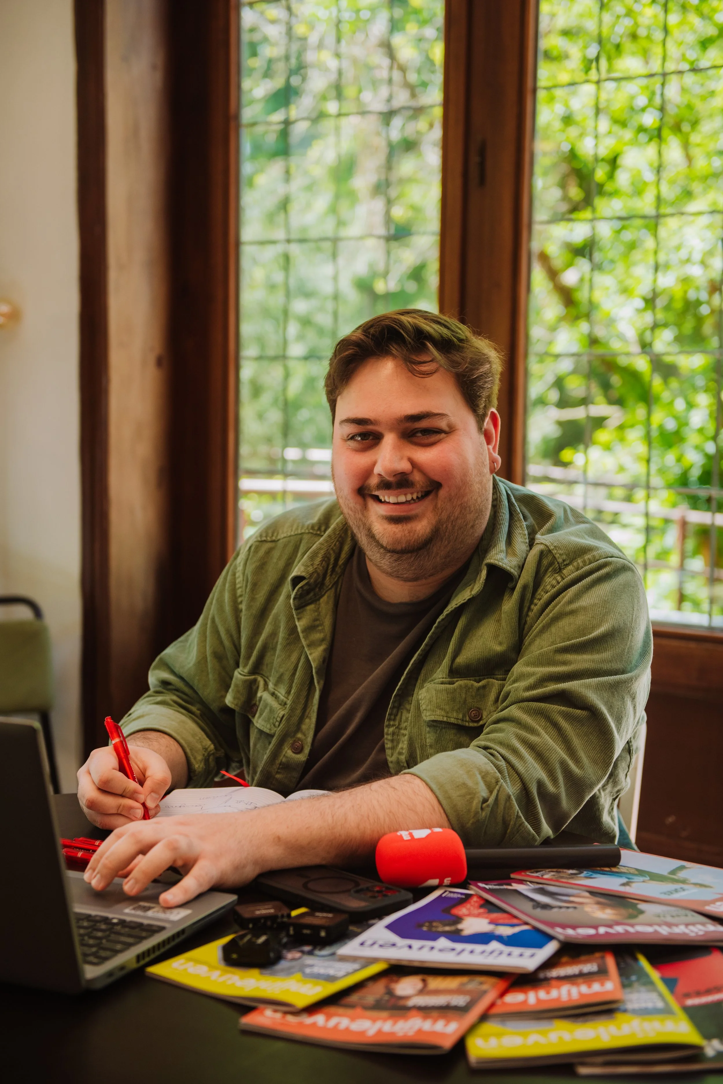 Een jonge heer aan een tafel met een computer, magazines en een microfoon