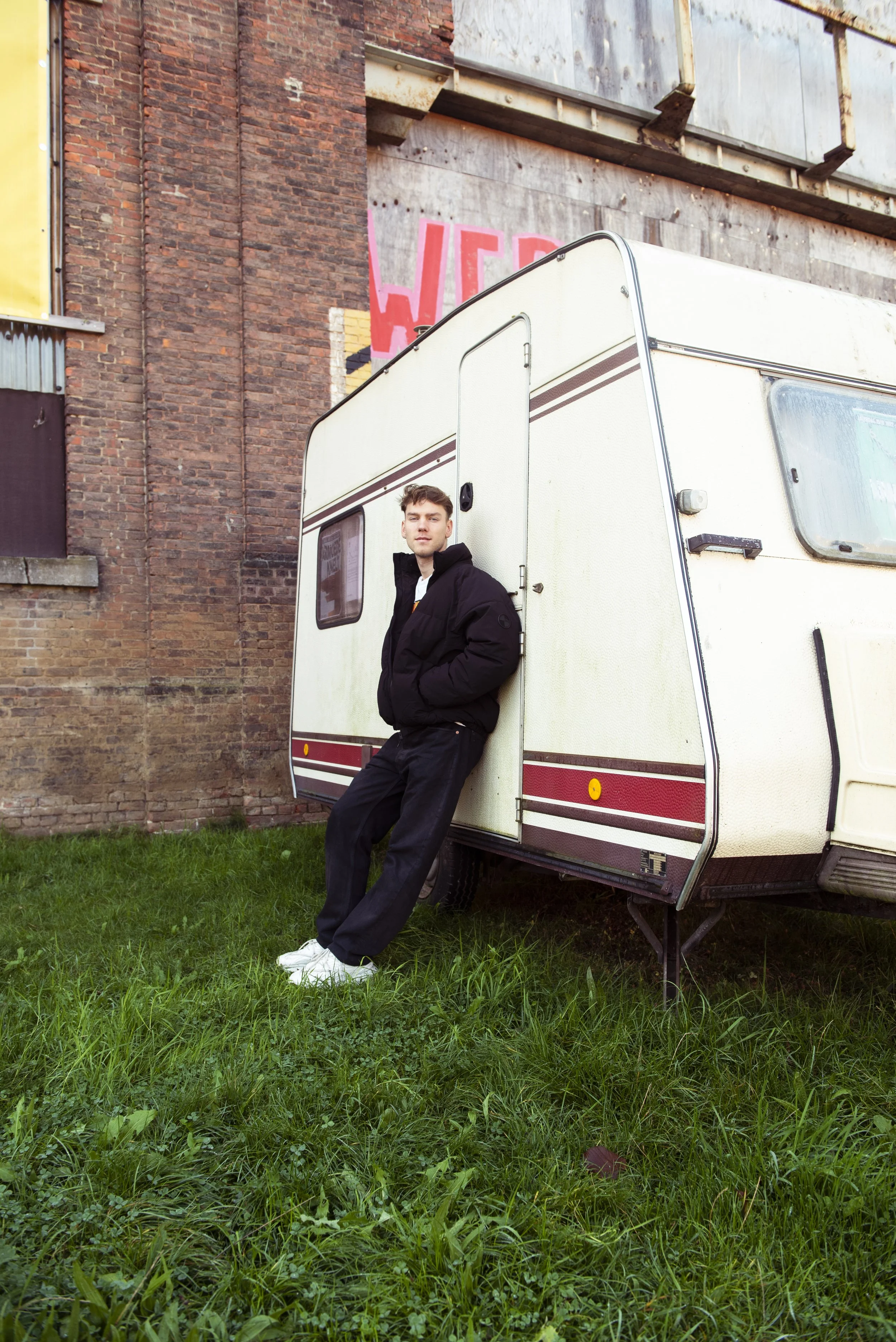 Frisse jonge man leunt tegen een witte caravan op een grasveld, achter een rode bakstenen muur met graffiti bevestiging.