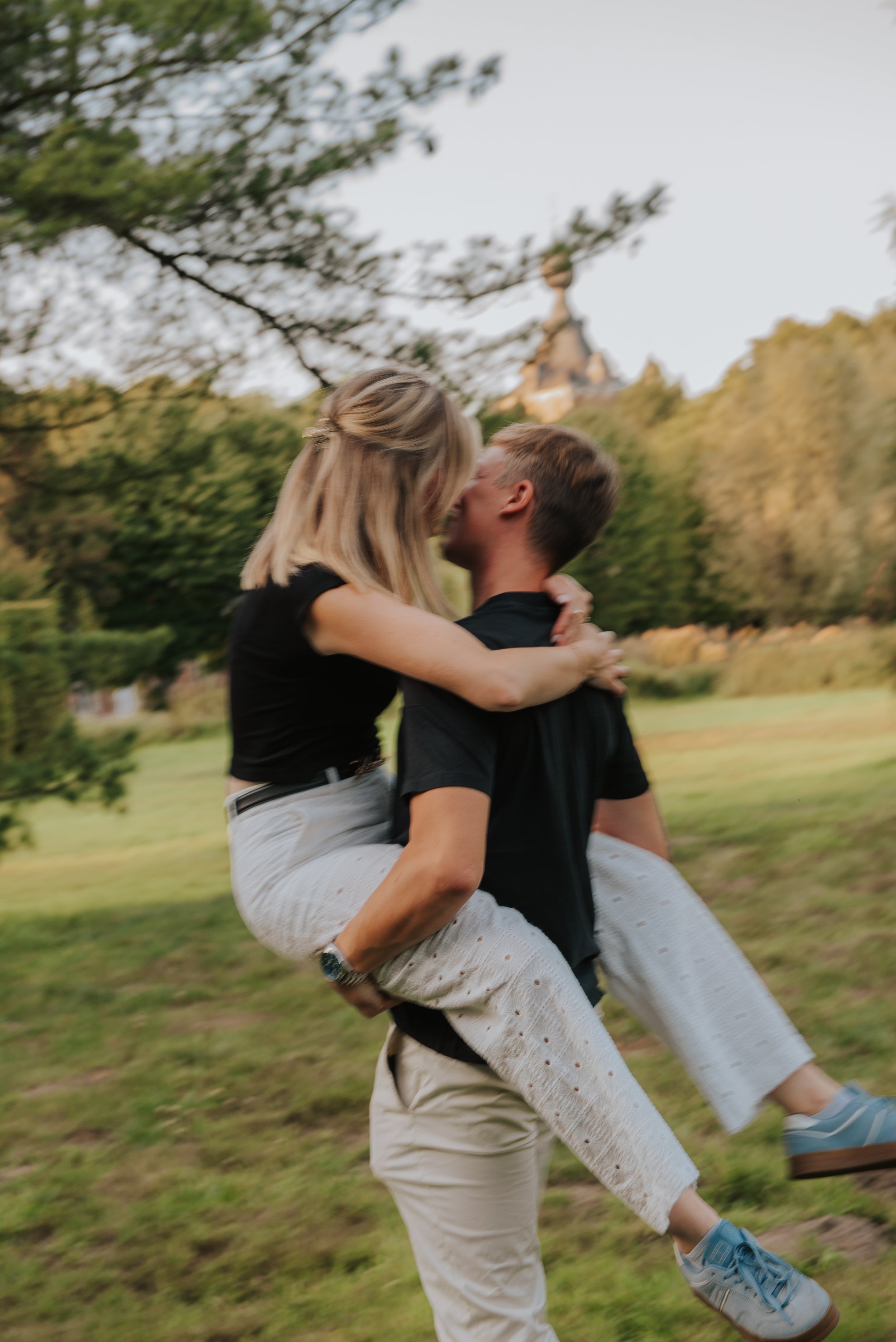 Een stel dat iemand op zijn rug draagt terwijl ze elkaar kussen in een park, met bomen en een gebouw op de achtergrond.