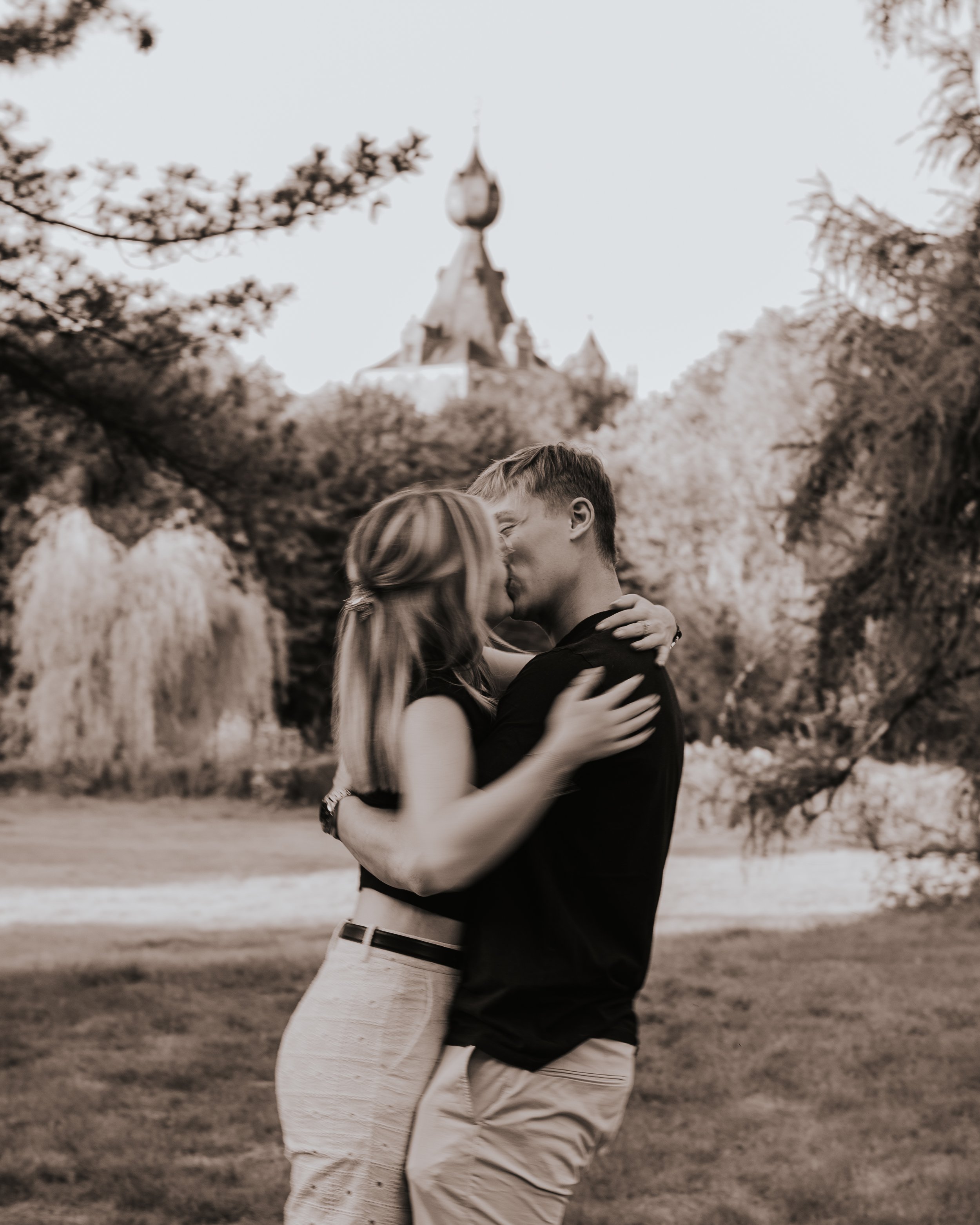 Een paar kust elkaar in een park met een kasteel op de achtergrond, in zwart-wit.