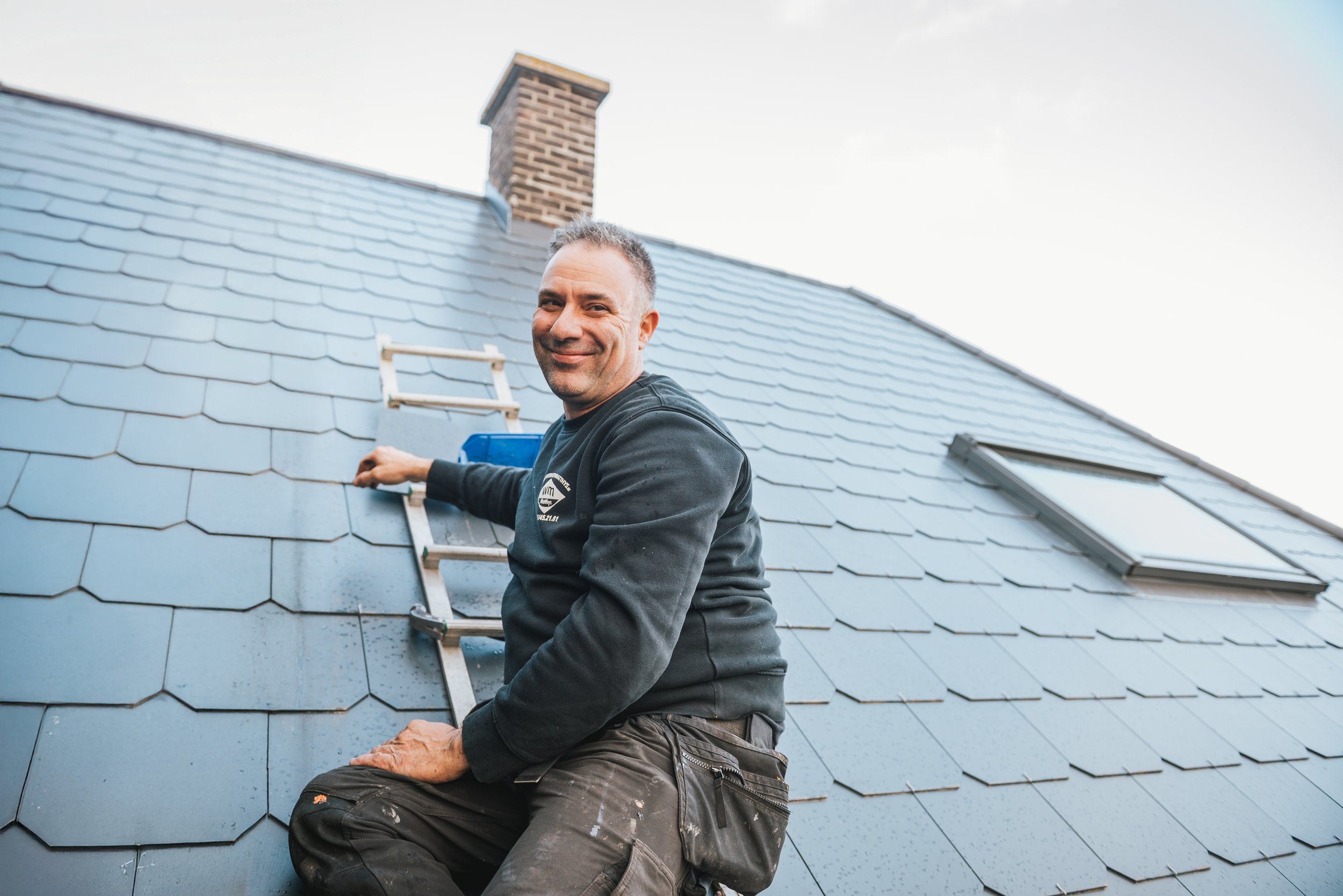 Man op het dak van een huis met nieuwe dakpannen, lachend naar de camera.