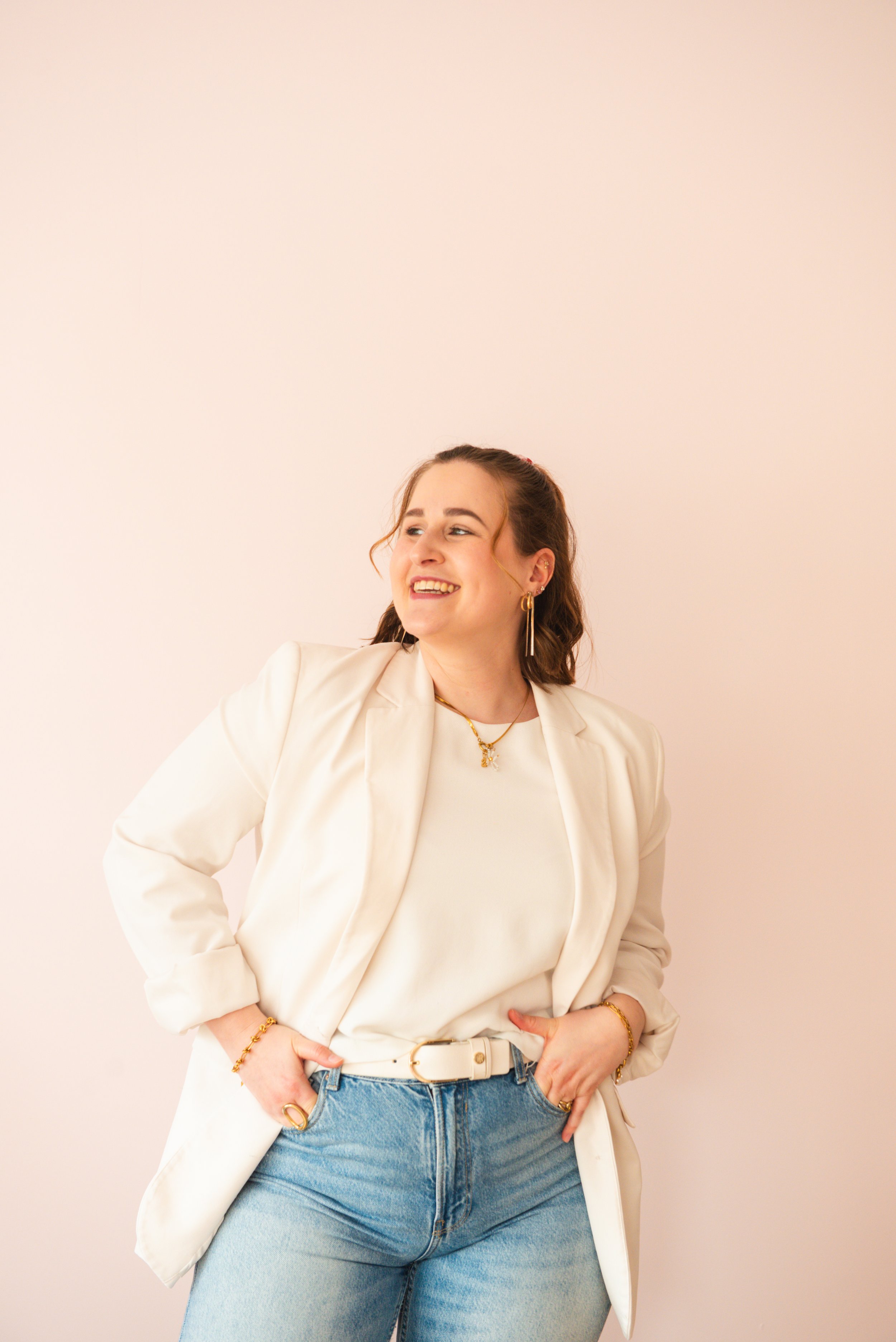 Vrouw met bruin haar lachend, witte blazer en jeans, poseert lachend tegen een lichte roze muur.