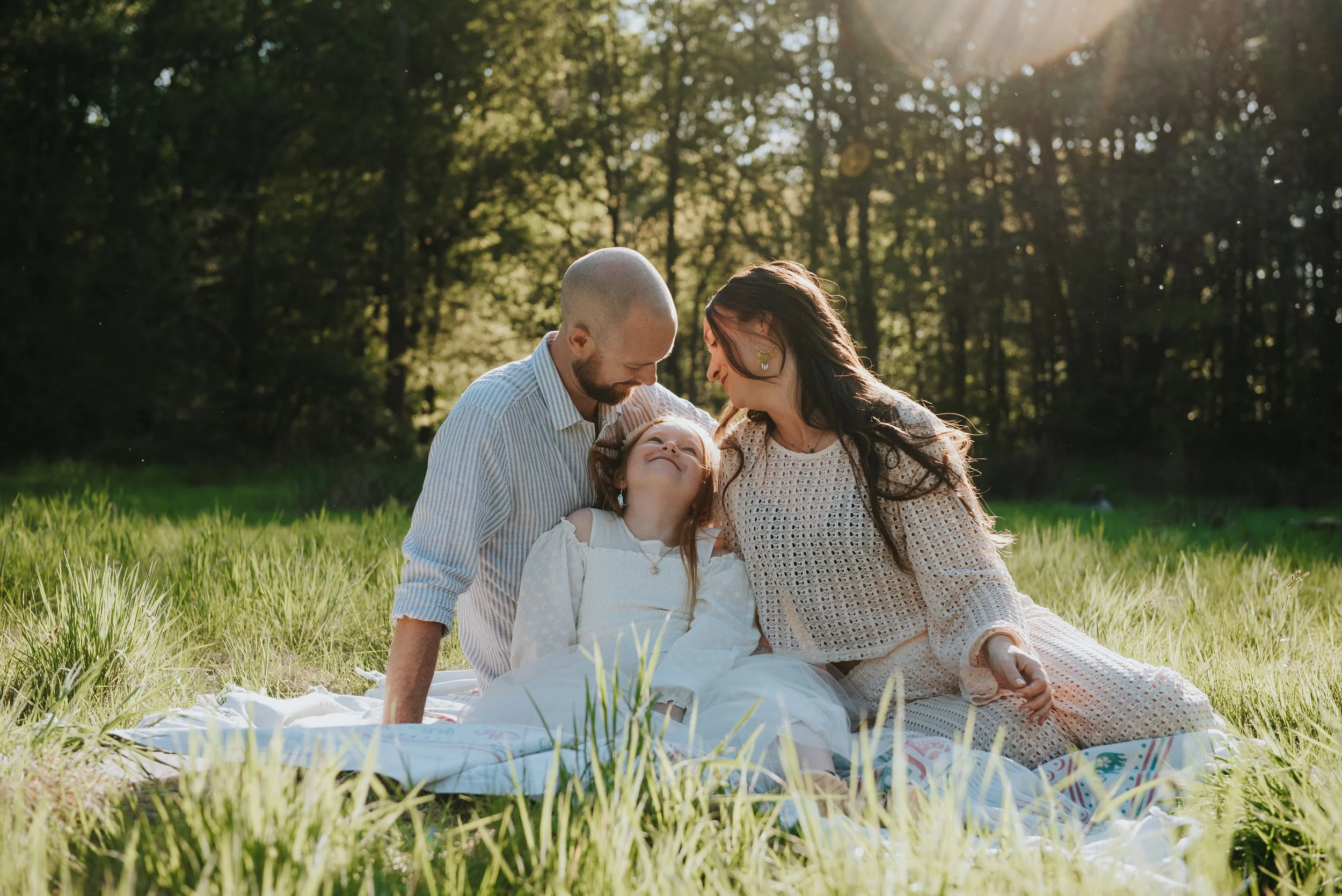 Gezin die samen op een grasveld in de zon zit, met een bos in de achtergrond, en elkaar liefkozend.