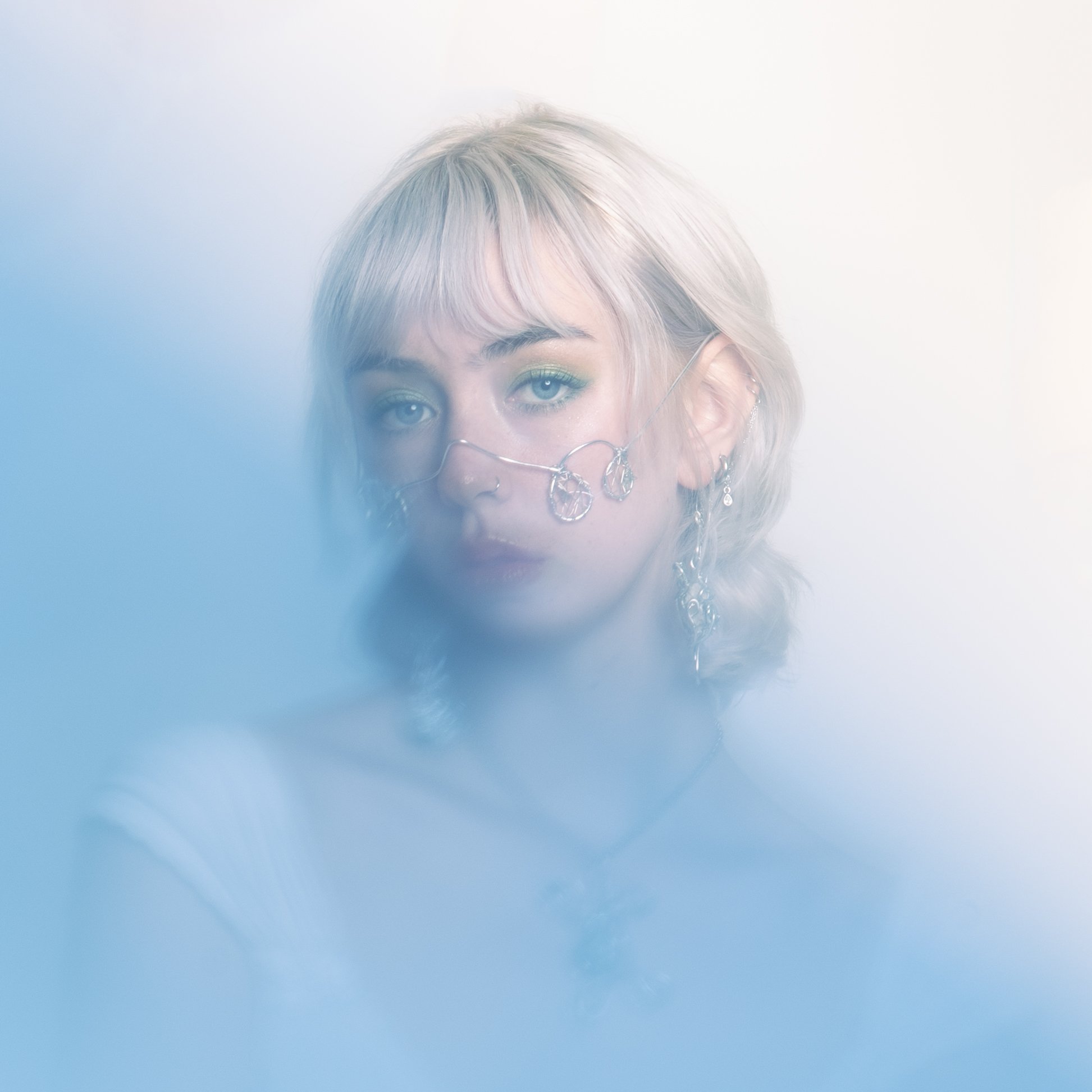 Portret van een jonge vrouw met licht blond haar en blauwe ogen, versierd met kettingen en oorbellen, omgeven door een wazige, blauwe en witte achtergrond.