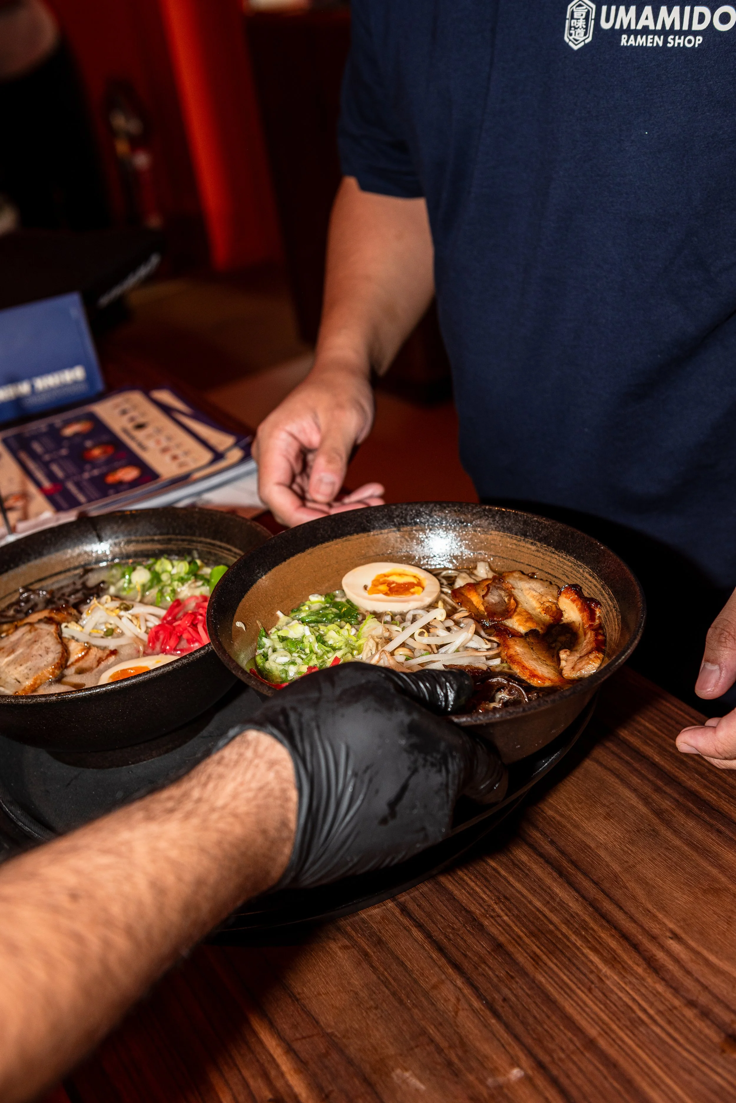 Een persoon serveert ramen in een restaurant, met een kom gevuld met noodles, een gekookt ei, varkensvlees en groenten.