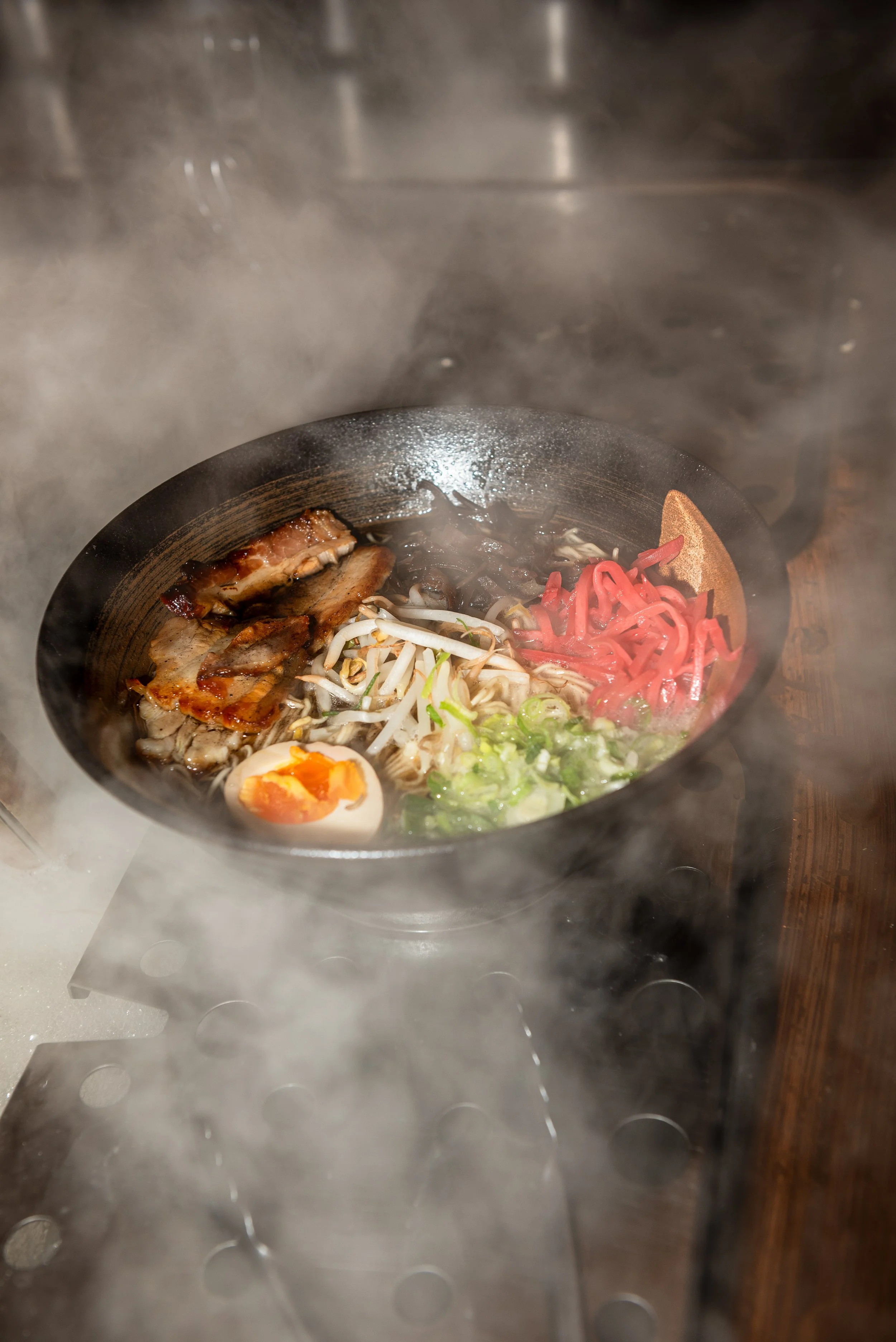 Een bowl ramen met vlees, een zachtgekookt ei, gesneden groenten, en noedels, gestoomd in een rokerige omgeving.