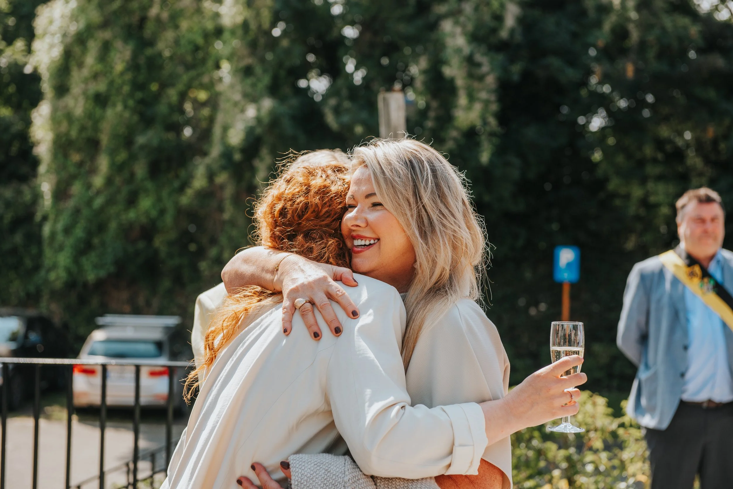 Twee vrouwen omhelzen elkaar vrolijk tijdens een buitenfeest, met een glas champagne in de hand. Op de achtergrond een man in een grijze jas met gele sjerp, groene bomen en een blauwe parkeersignaal.