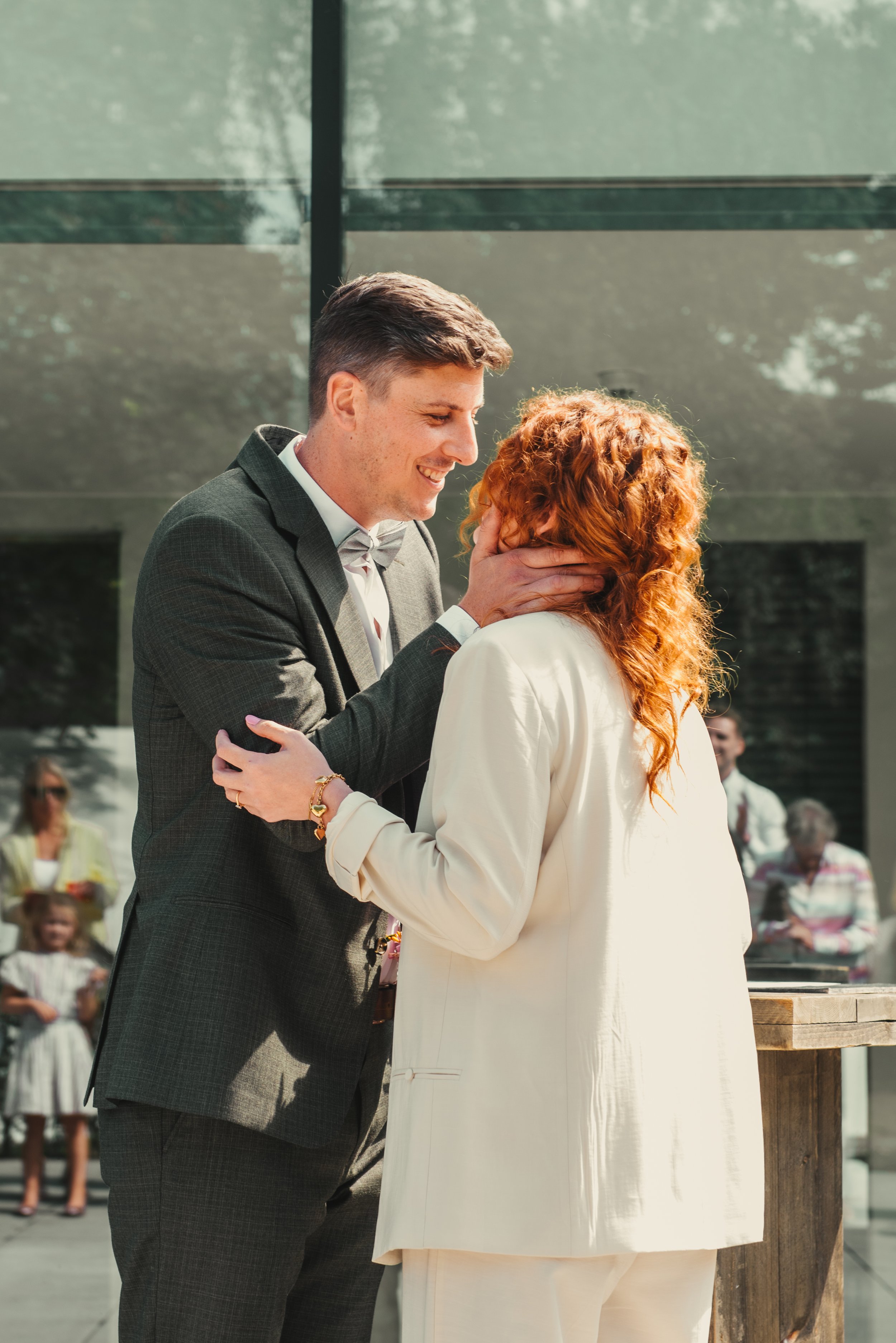 Man en vrouw in witte en grijze kleding houden elkaar vast en kussen op een buitenlocatie tijdens een bruiloft of ceremonie.