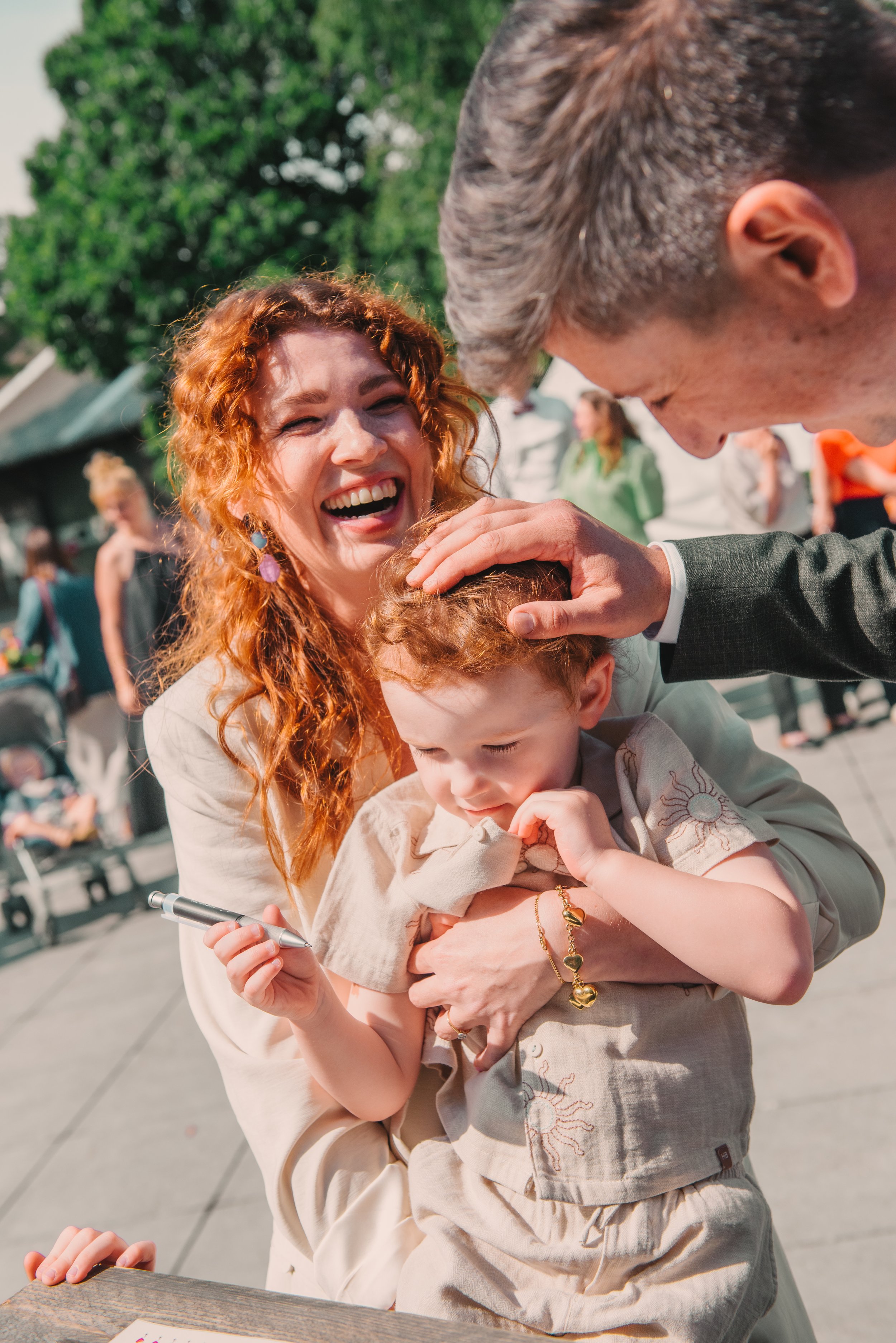 Een vrouw met rood haar lacht terwijl een man haar het hoofd aait en een jongen vastpakt die een pen vasthoudt, op een zonnig dag buiten.