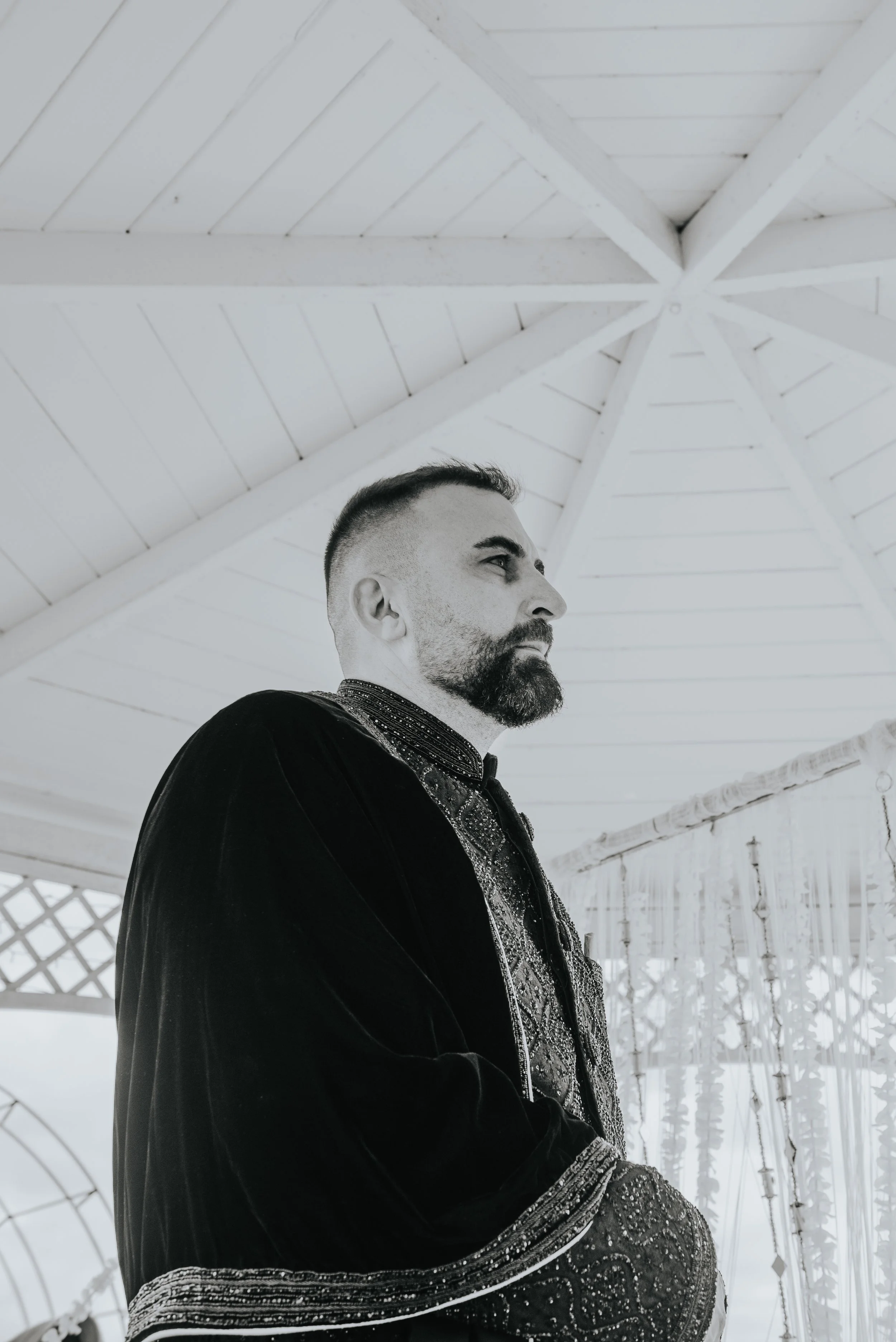 Een man met een baard en kort haar, gekleed in een zwarte, rijkelijk versierde jas, staat onder een witte, houten gazebo met een decoratie van zwevende ornamenten aan de rechterkant.