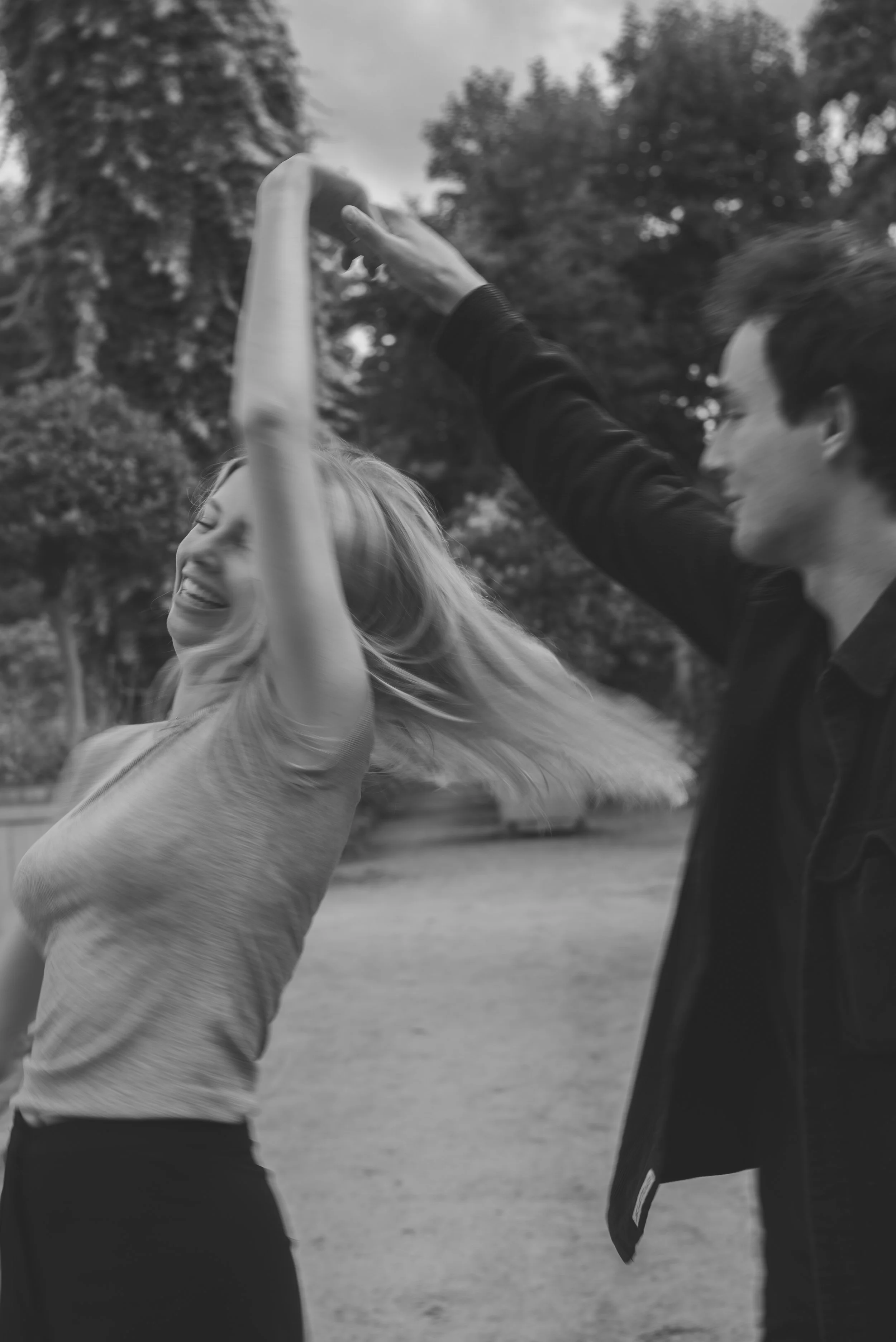 Een lachende vrouw met lang haar danst terwijl een man haar een hoedje geeft in een park op een bewolkte dag.