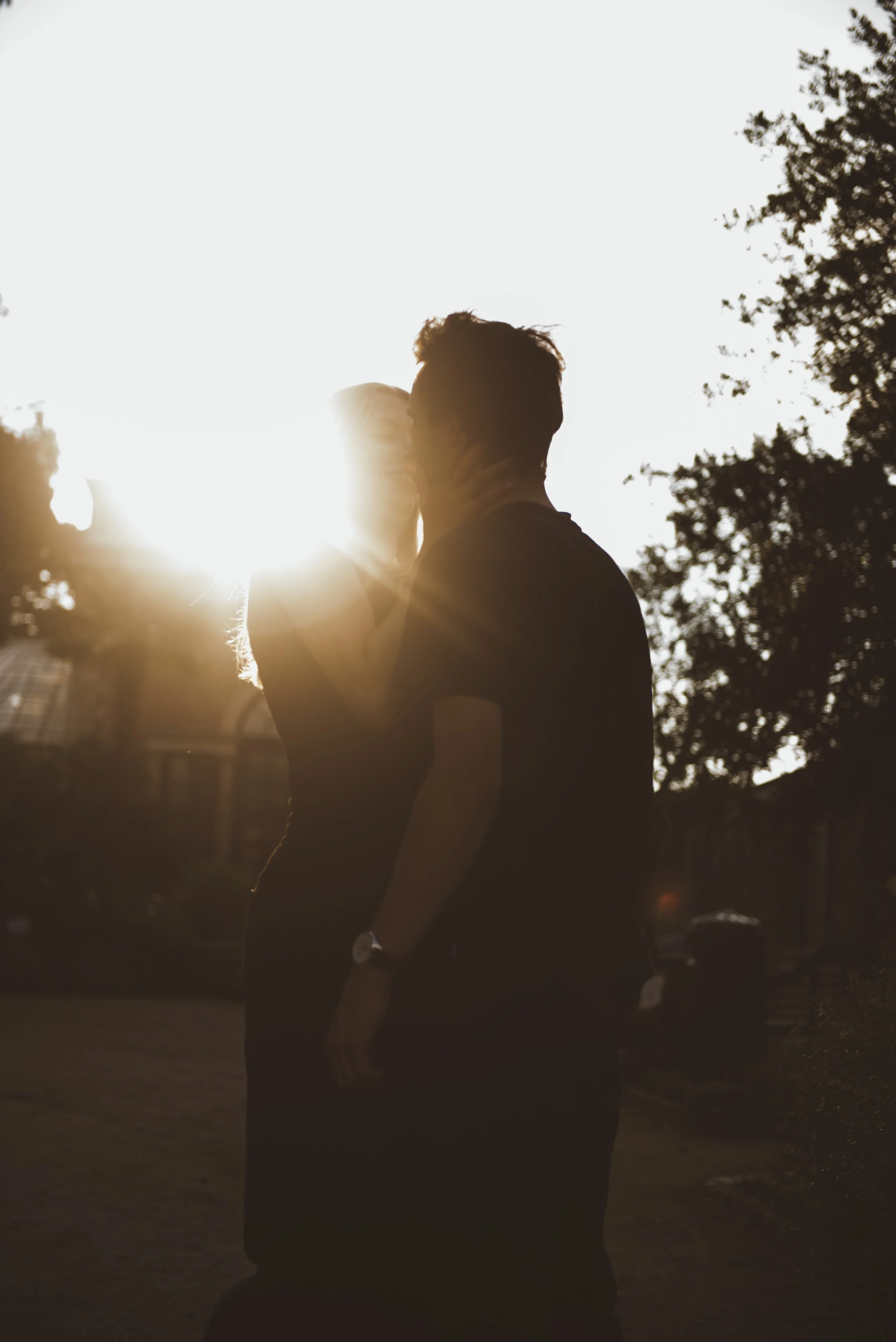 Koppel die elkaar kust tijdens zonsondergang, silhouet zichtbaar, omgeven door bomen en een rustige omgeving.