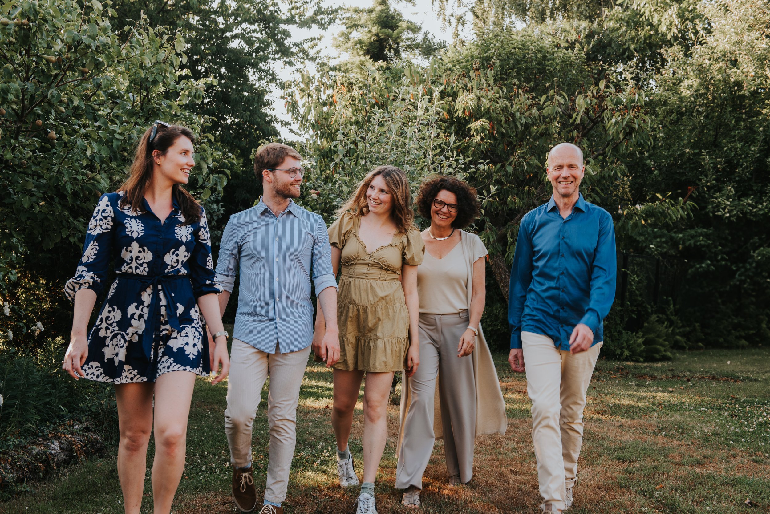 Vijf mensen wandelen samen in een tuin met groene bomen, lachen en praten.