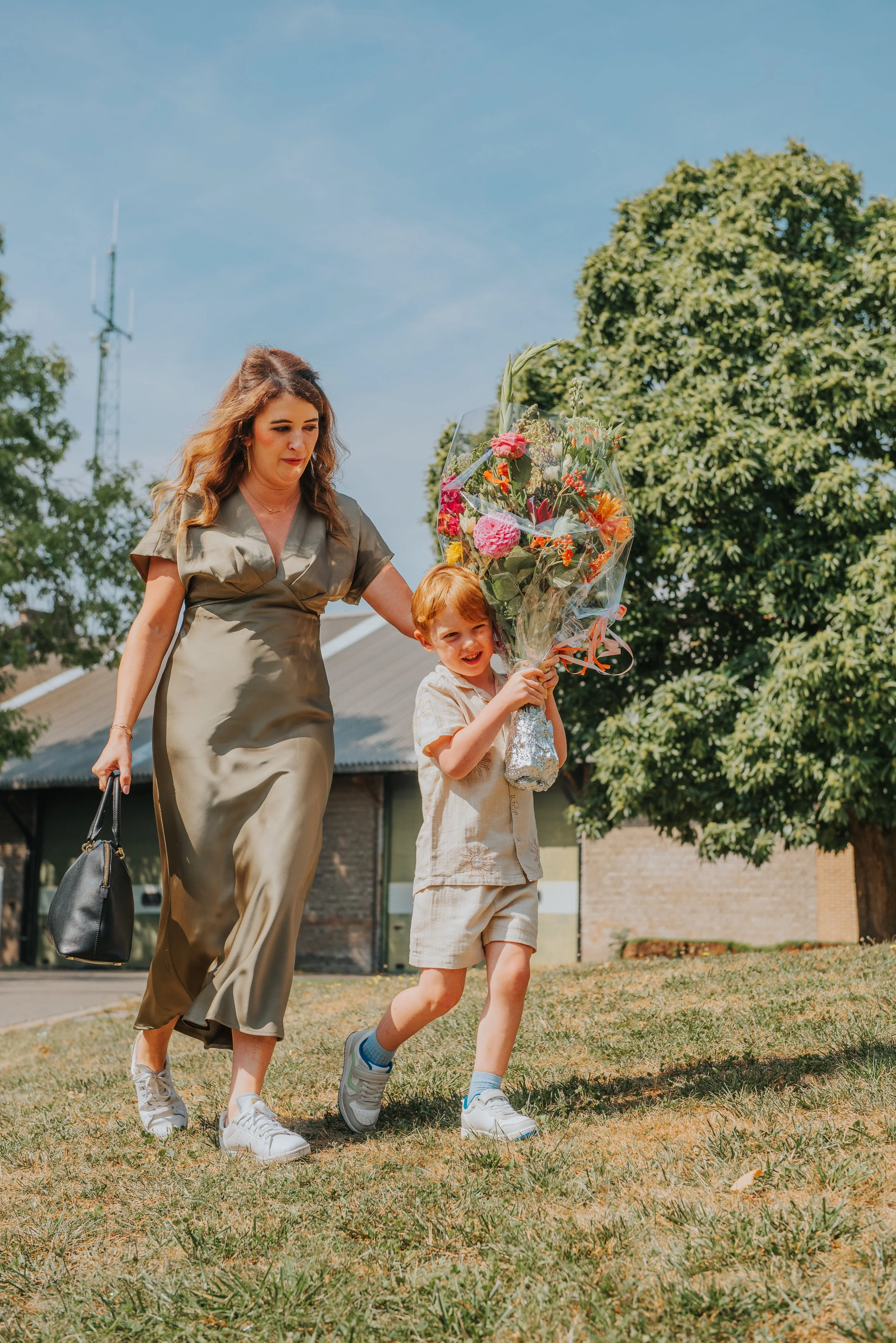 Een vrouw en een jong kind lopen samen op gras, de vrouw houdt de arm van het kind vast en geeft hem een bos bloemen.