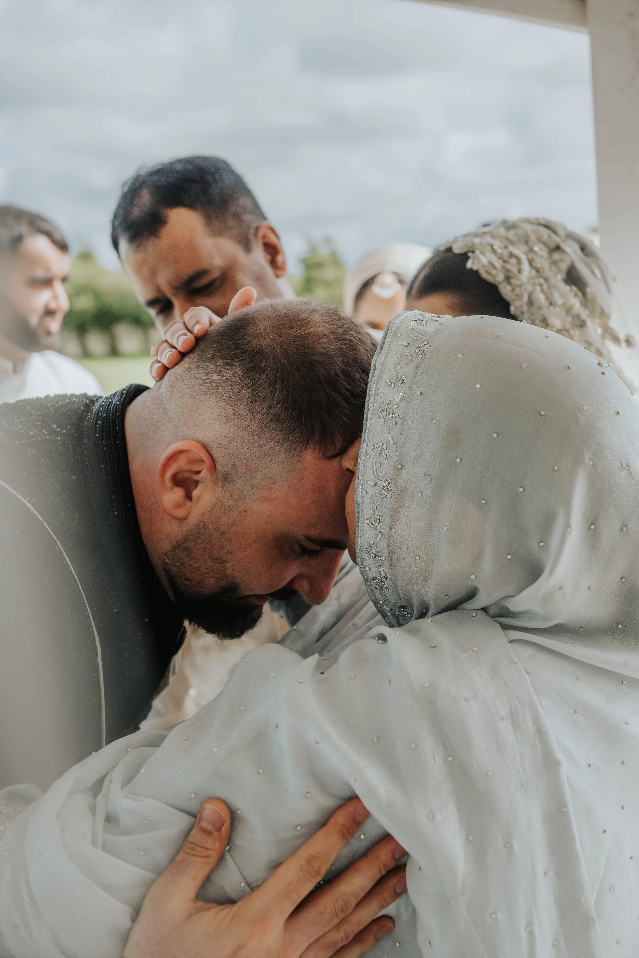 Een huwelijksceremonie waarbij een man en een vrouw elkaar in de ogen kijken terwijl ze hun voorhoofden tegen elkaar duwen, omringd door getuigen en familie op een heldere dag.