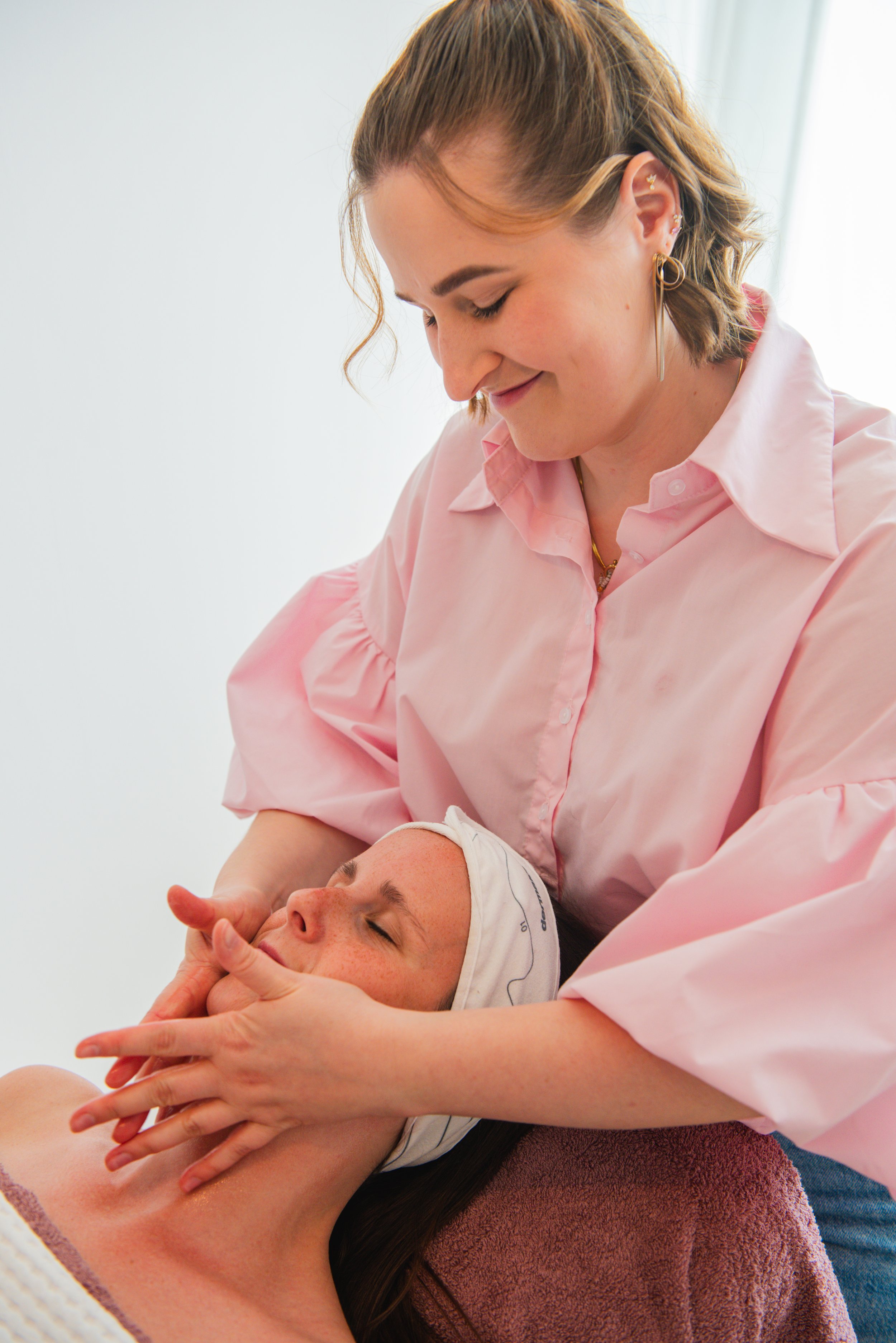 Een vrouw die haar gezicht verzorgt bij een schoonheidsbehandelingen of massage, liggend op een bank met een handdoek om haar lichaam, terwijl een beautyspecialiste haar gezicht masseert.
