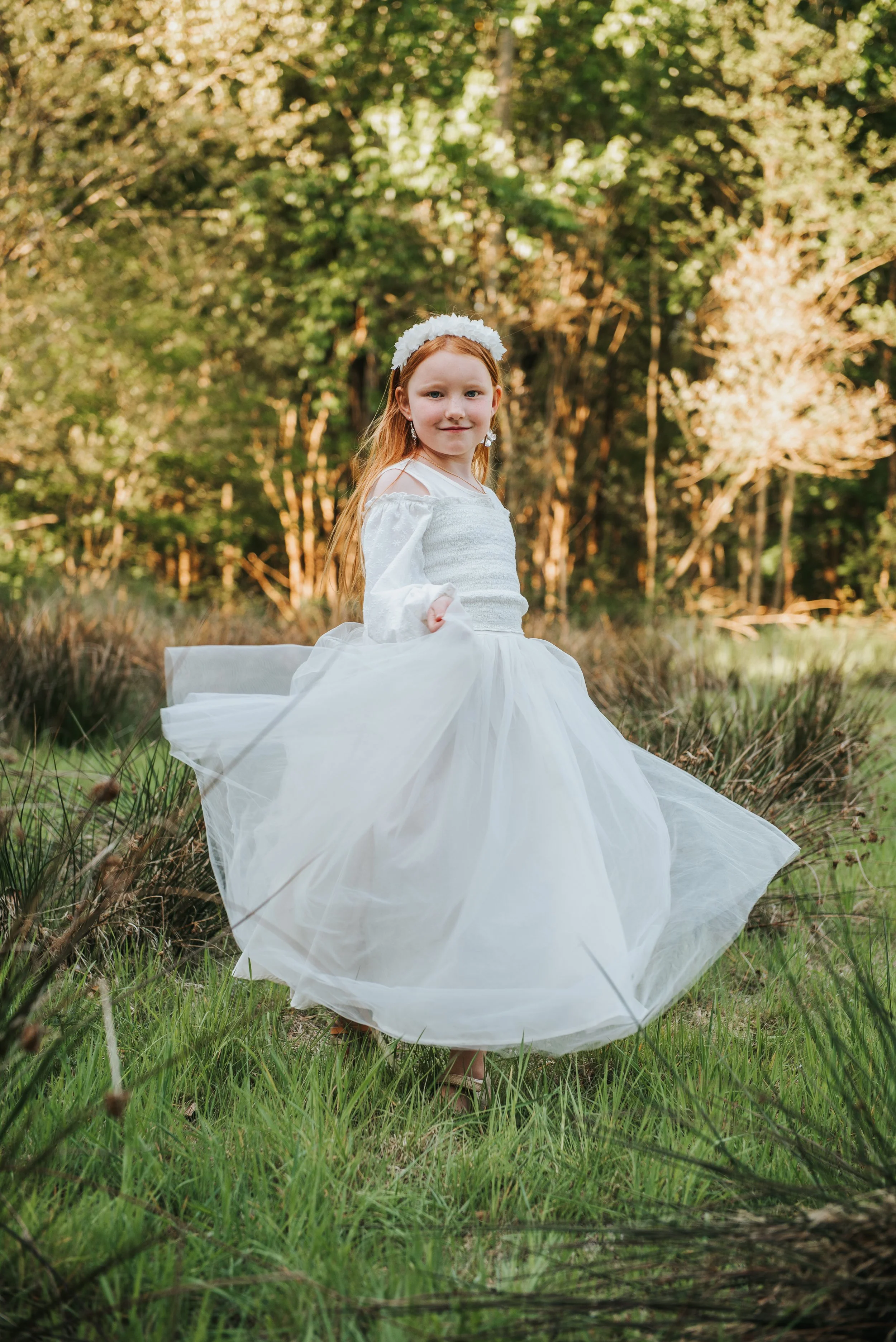 Een jong meisje in een witte jurkj over een grasveld in een bos, omringd door groene bomen, met een witte bloemenkroon in haar haar.