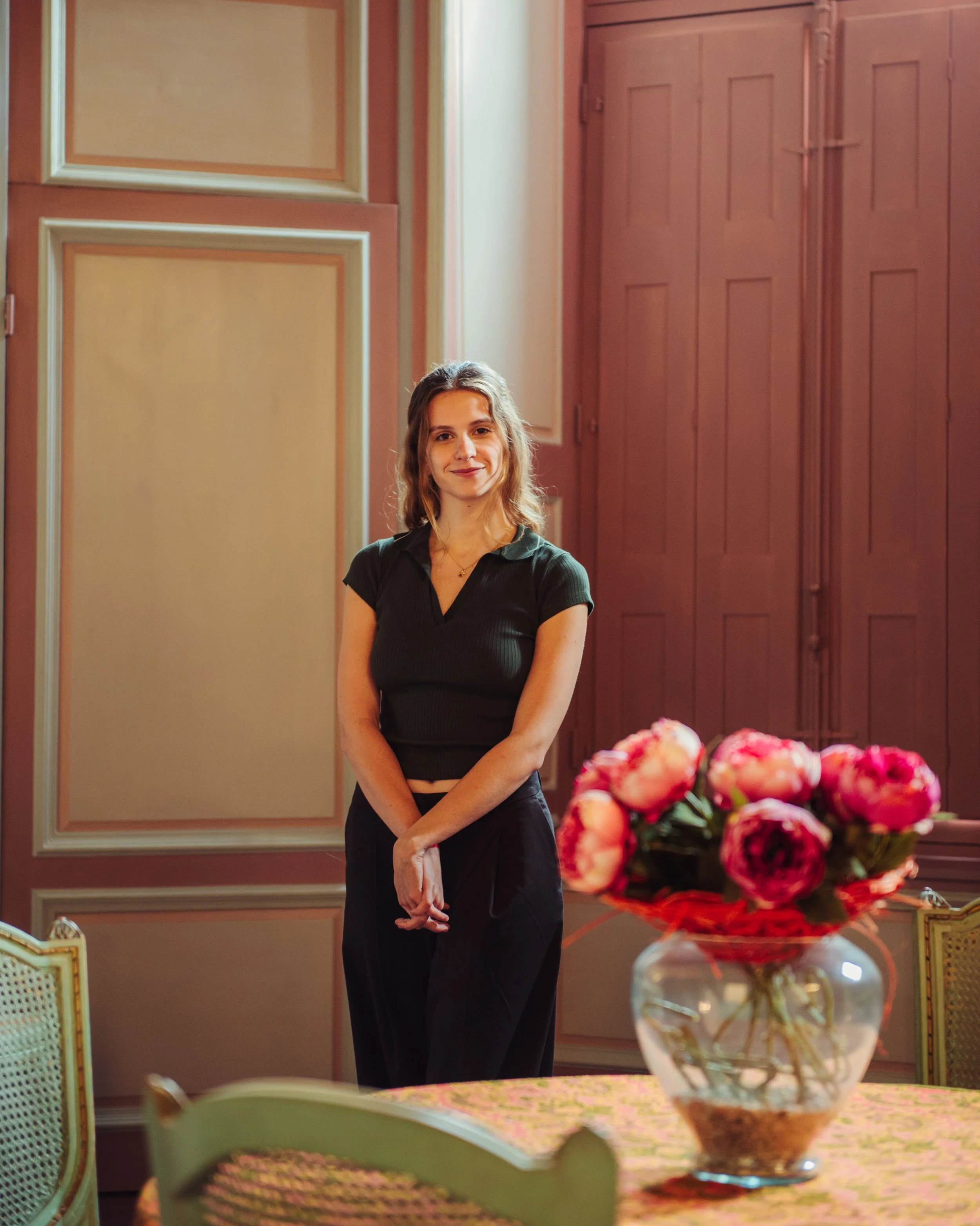 Een jonge vrouw staat in een elegante kamer met houten panelen op de muur, met een grote vaas met roze bloemen op een gedekte tafel voor haar.