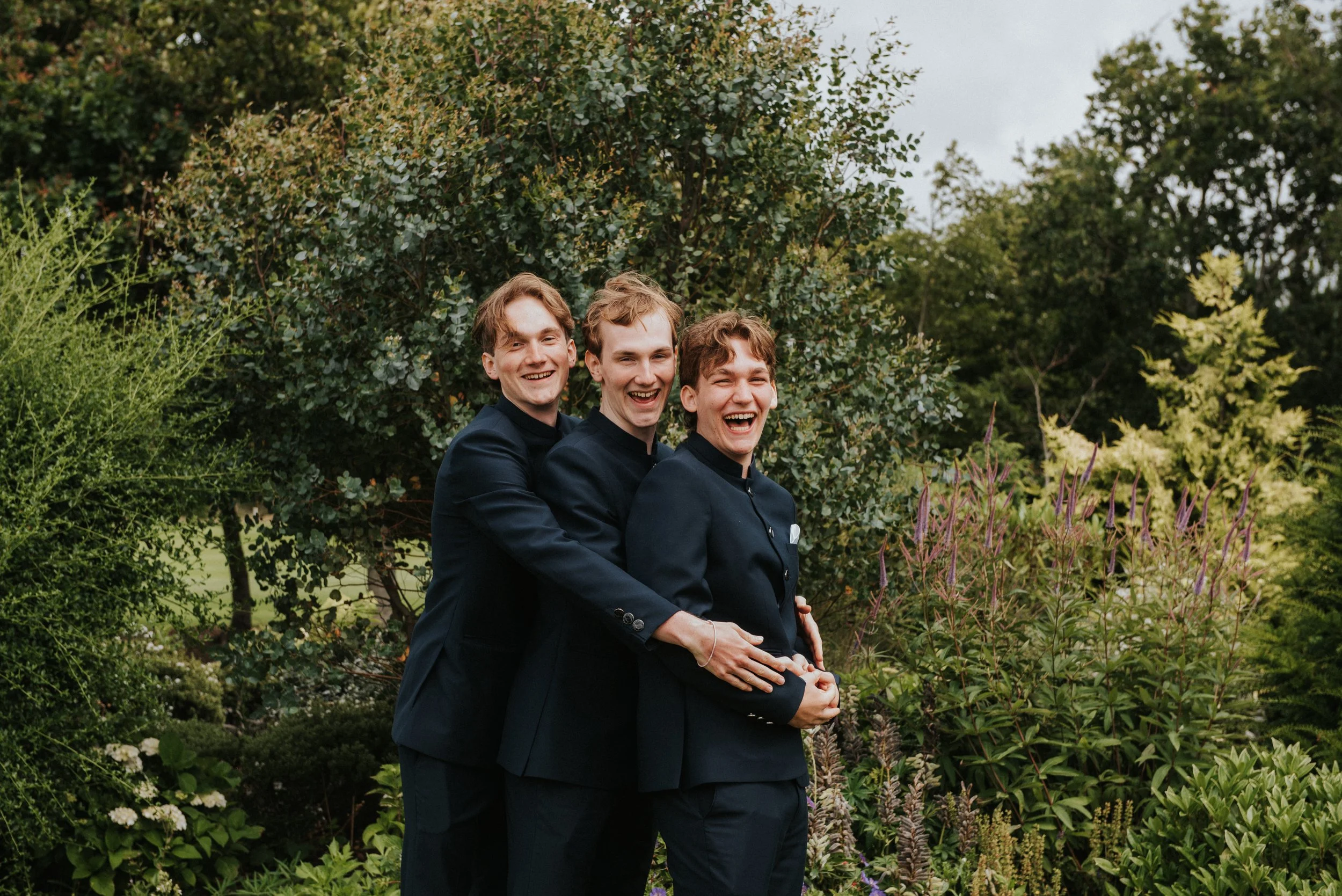Drei jonge mannen in zwarte pakken lachen en houden elkaar vast in een tuin met groene struiken en bomen.