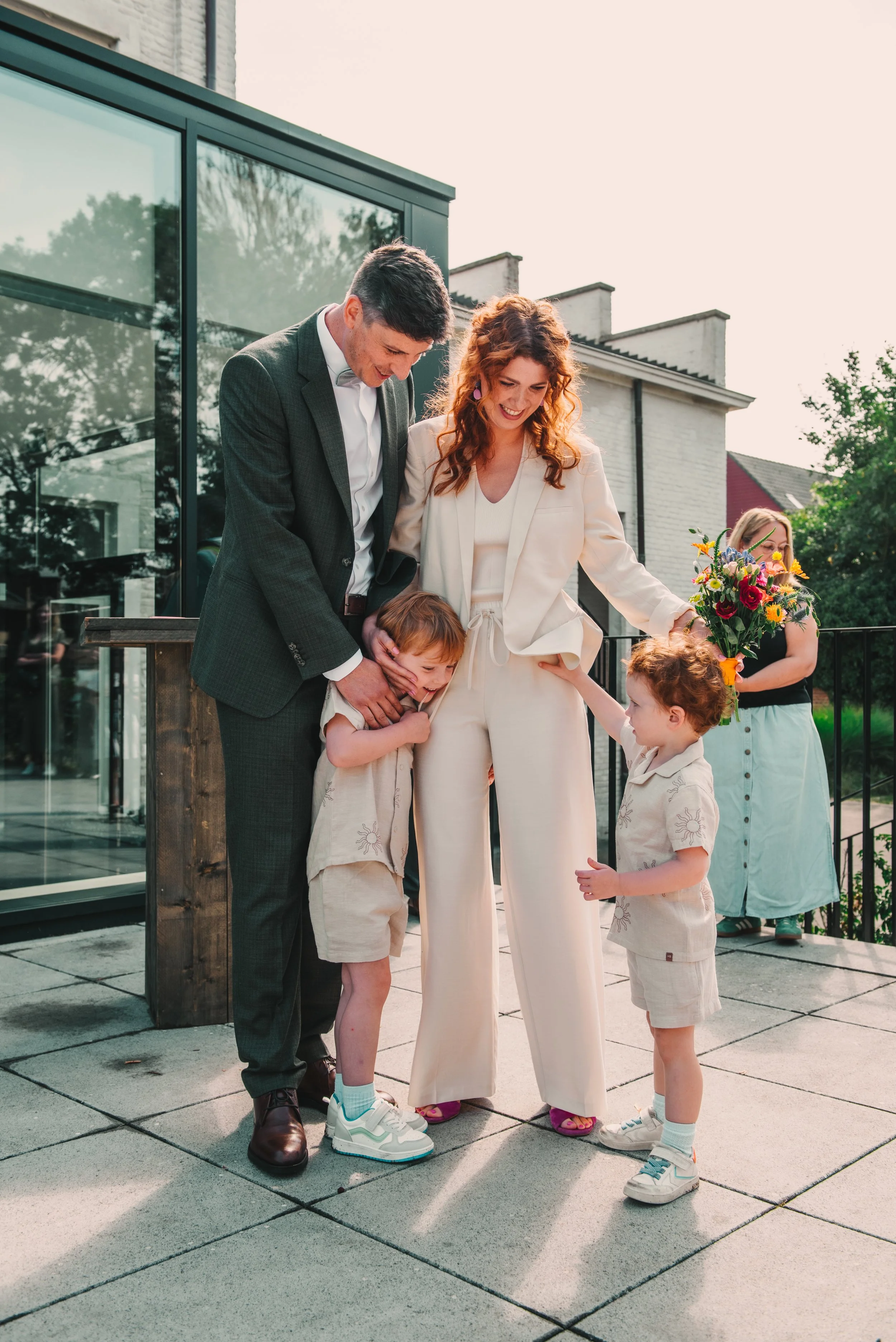 Een familie op een terras, lachend en knuffelend, met twee jonge kinderen en een vrouw met bloemen in haar hand.