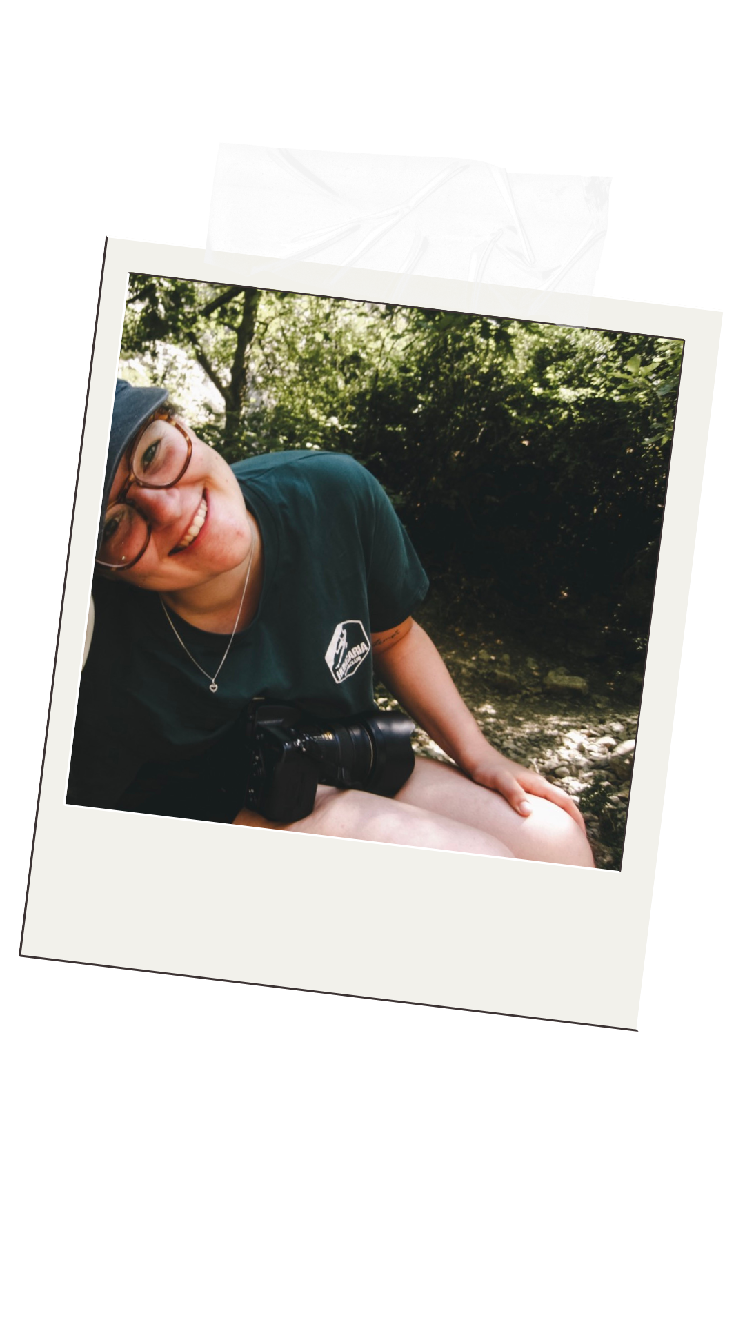Een glimlachende jonge vrouw met bril en pet, zittend bij een rivier in een bos, met een camera op haar schoot.