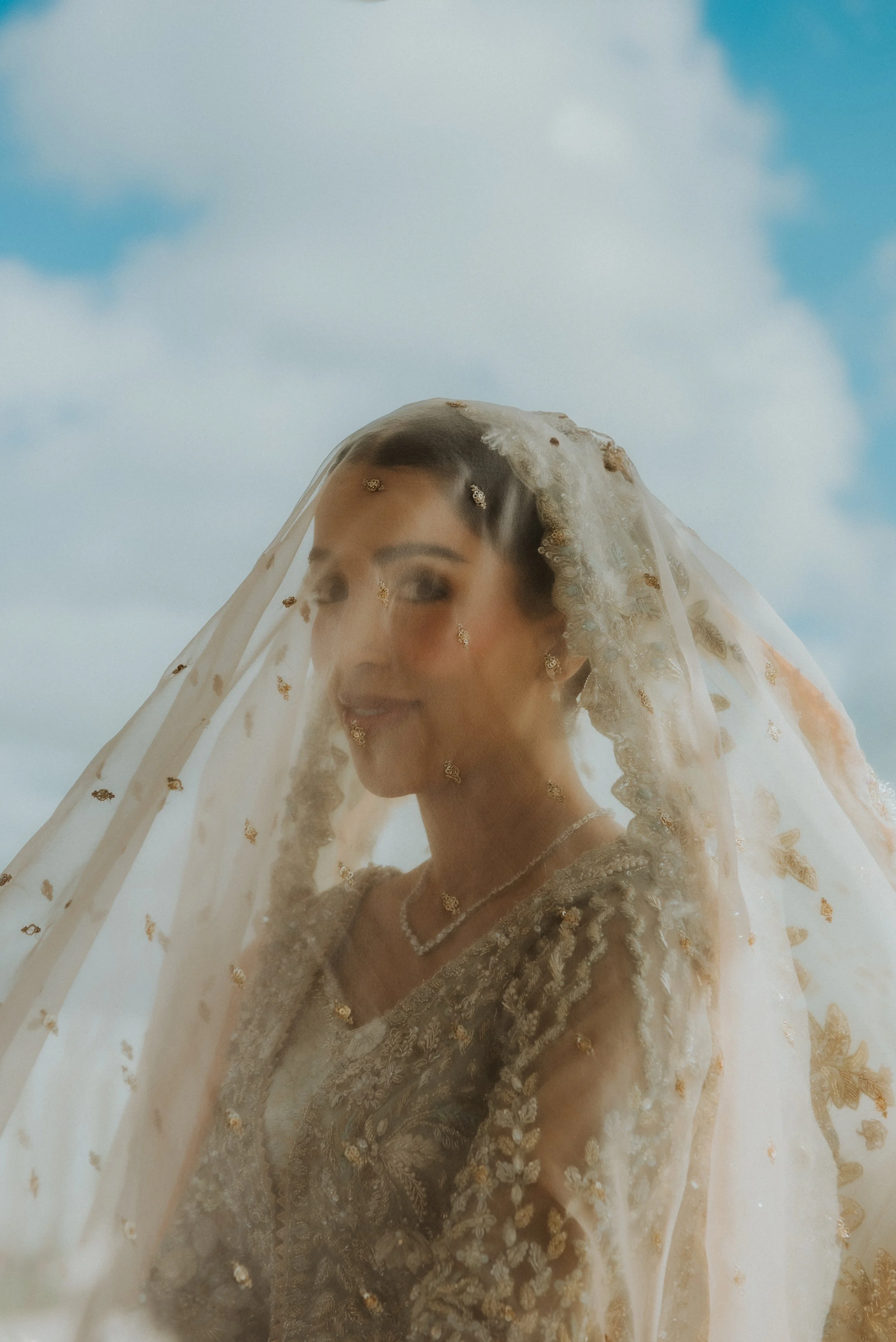 Een vrouw in een bruidsjurk, bedekt met een delicatieluchtige sluier met gouden details, die door de wind wordt opgevoerd, met een blauwe lucht en witte wolken op de achtergrond.