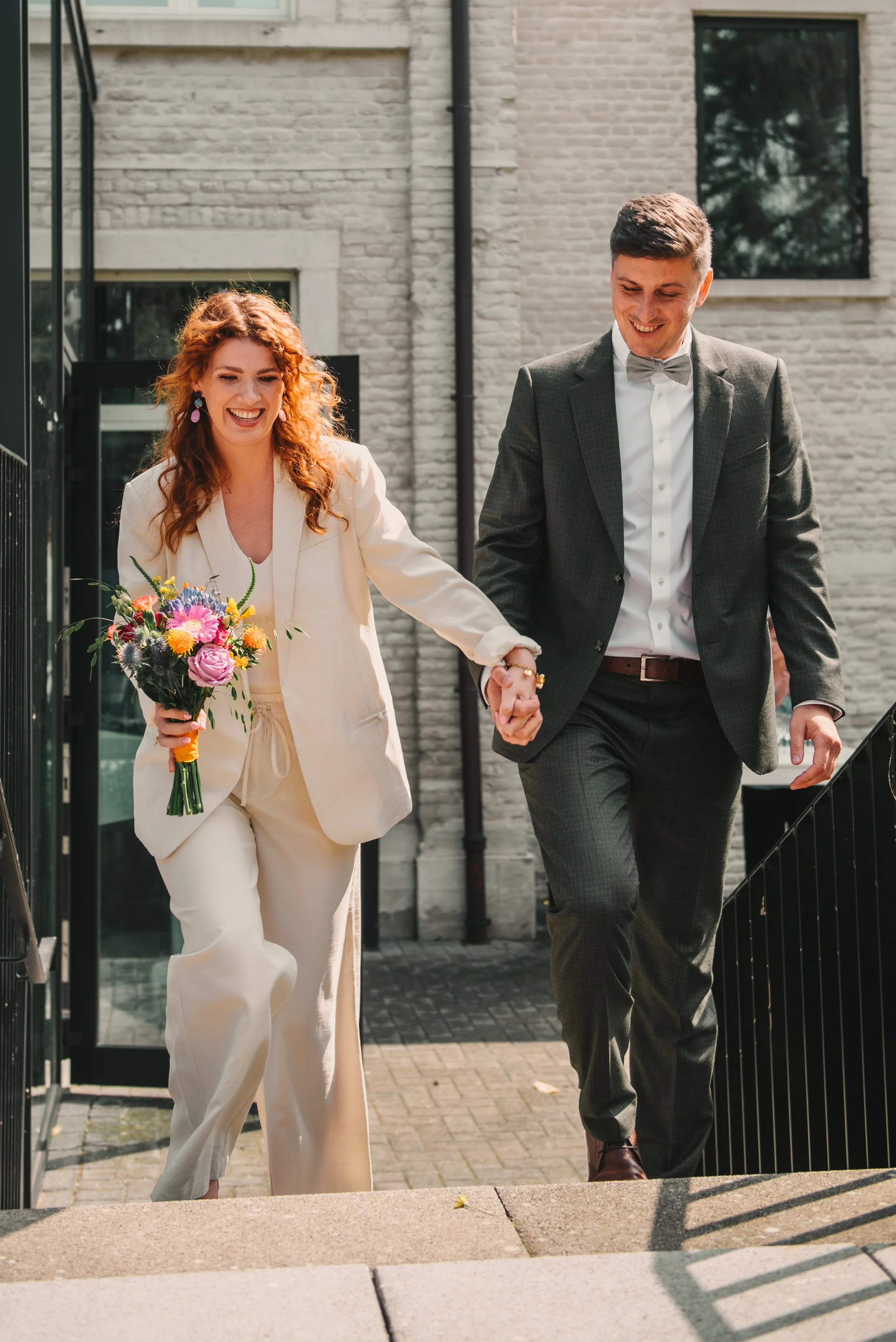 Twee mensen, een vrouw met een bloemenboeket en een man in een pak, lopen samen hand in hand de trap op terwijl ze lachen.
