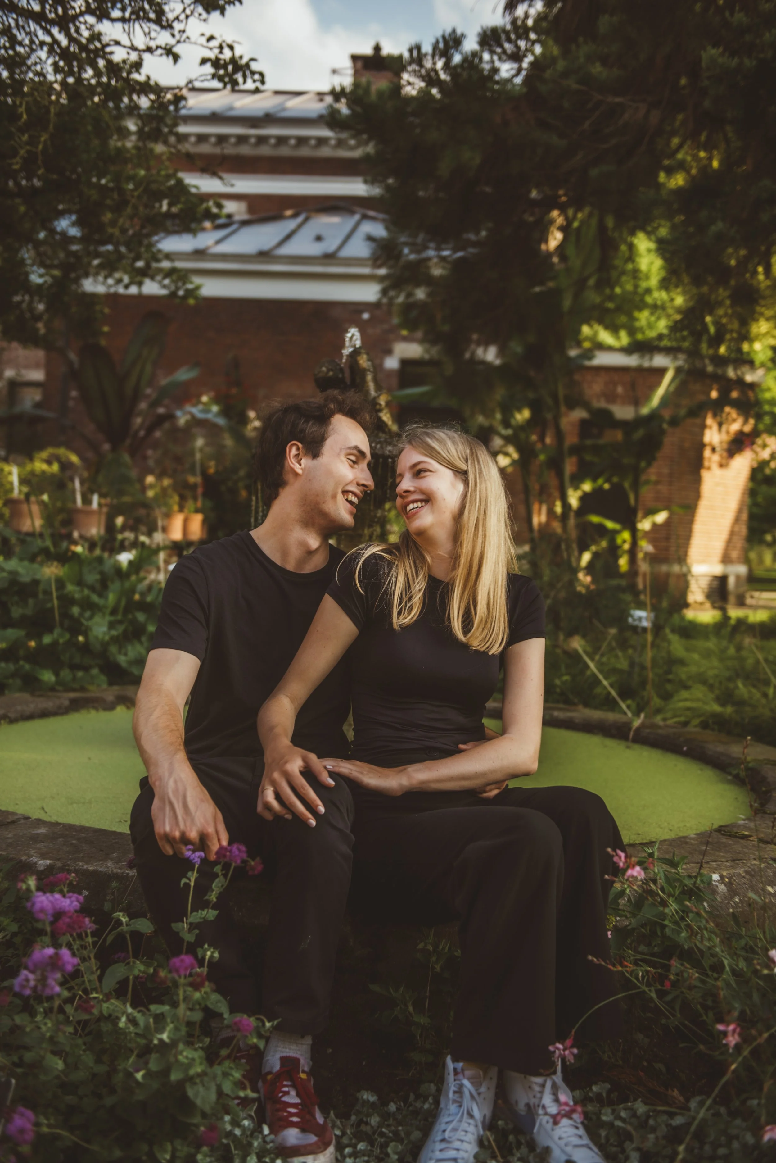 Een koppel zit samen op een bankje in een tuin, lachend en verliefd, omgeven door bloemen en groene planten.