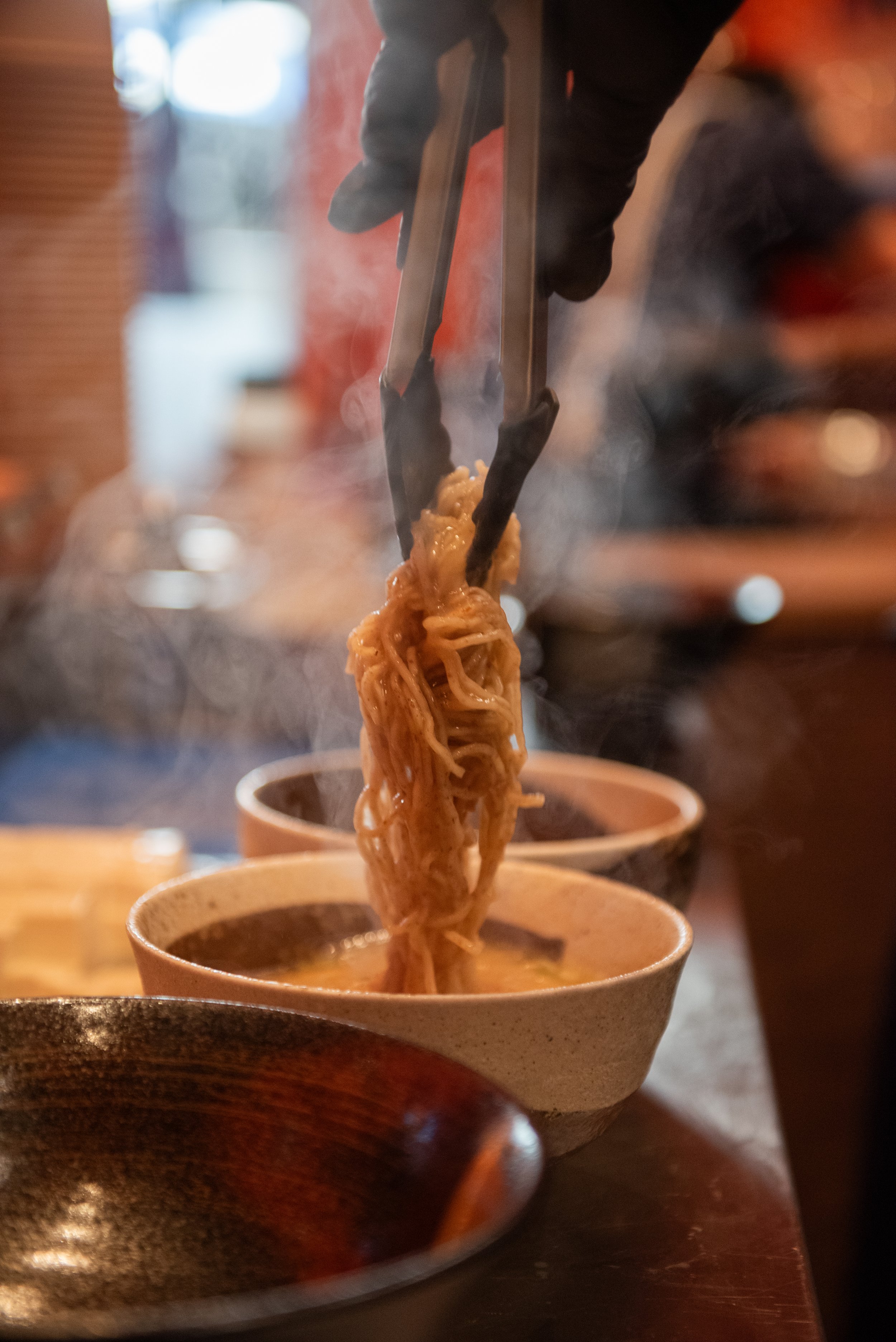 Persoon pakt een portie noedels met zwarte tangen boven een kom warme soep in een restaurant.