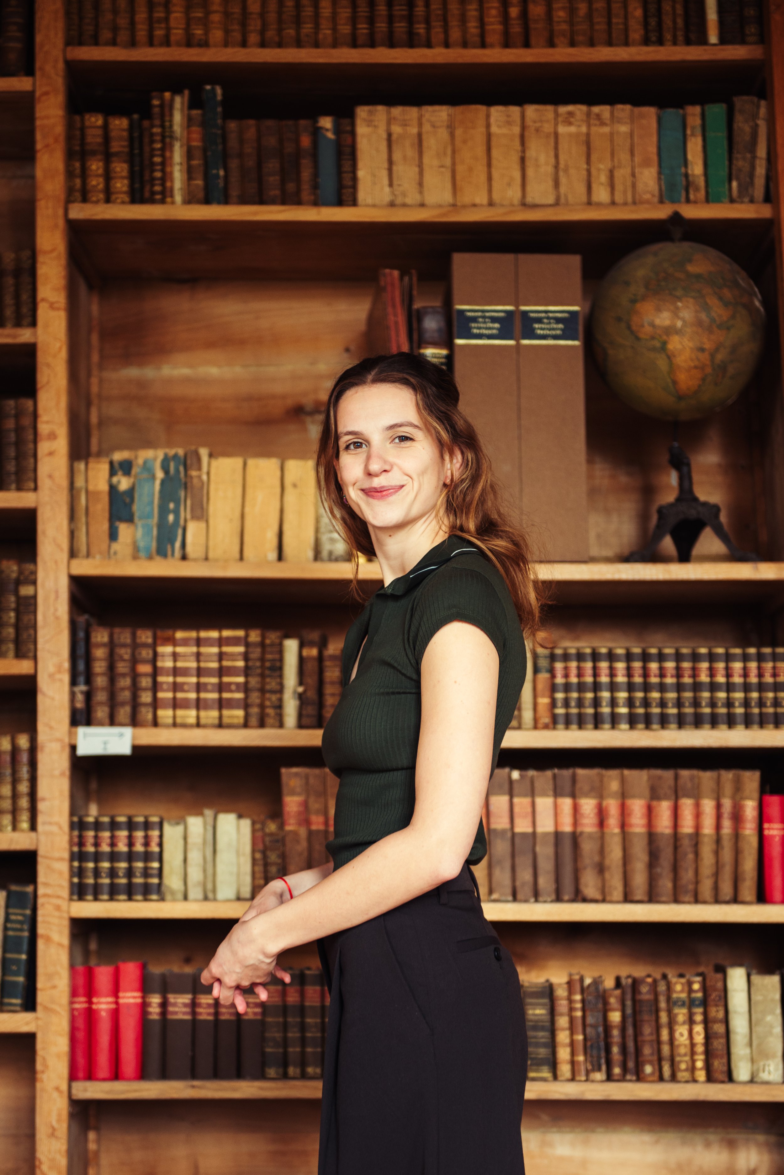 Vrouw met bruin haar in een groene blouse en zwarte broek poseert in een bibliotheek met houten boekenplanken, een wereldbol en oude boeken.