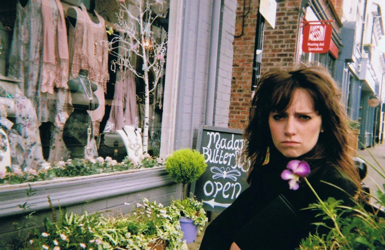 A woman with dark, shoulder-length hair and a serious expression standing outside a boutique clothing store with a sign that says "Madam Buttercup." The store window displays dresses, jewe, and a mannequin, with decorative plants and flowers outside.