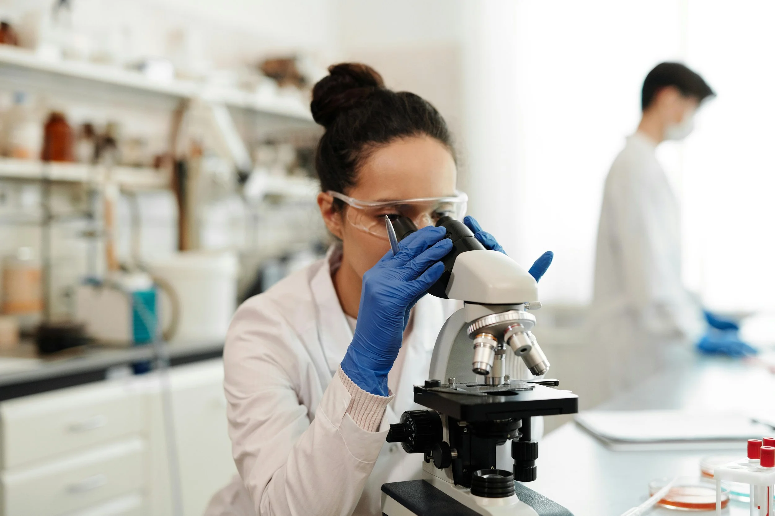 A female scientist in safety goggles and blue gloves looks through a microscope in a laboratory with another scientist in the background.