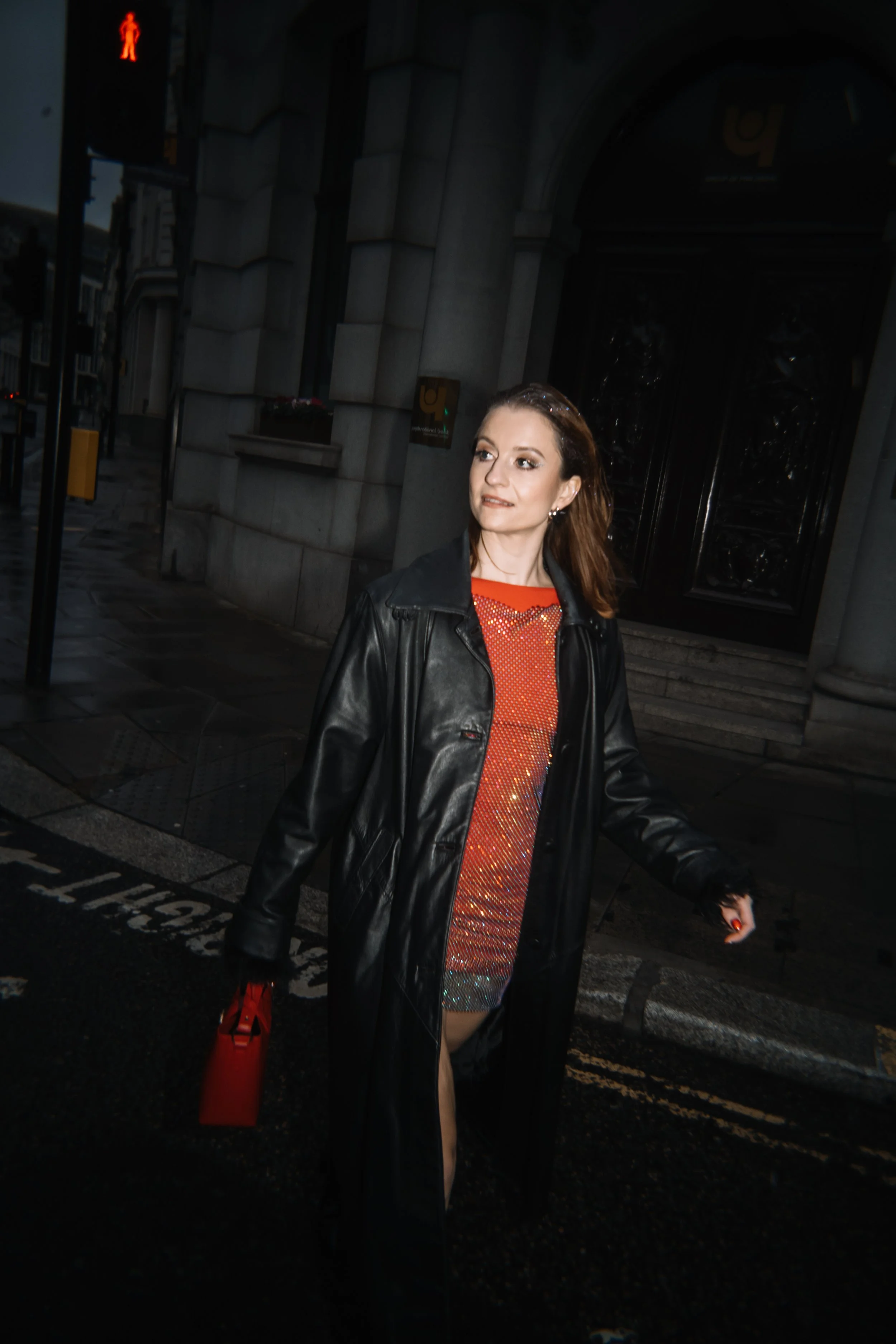 Woman walking at night, wearing a black leather jacket and a red and orange sequin dress, with a red bag and holding a phone, in an urban street scene.