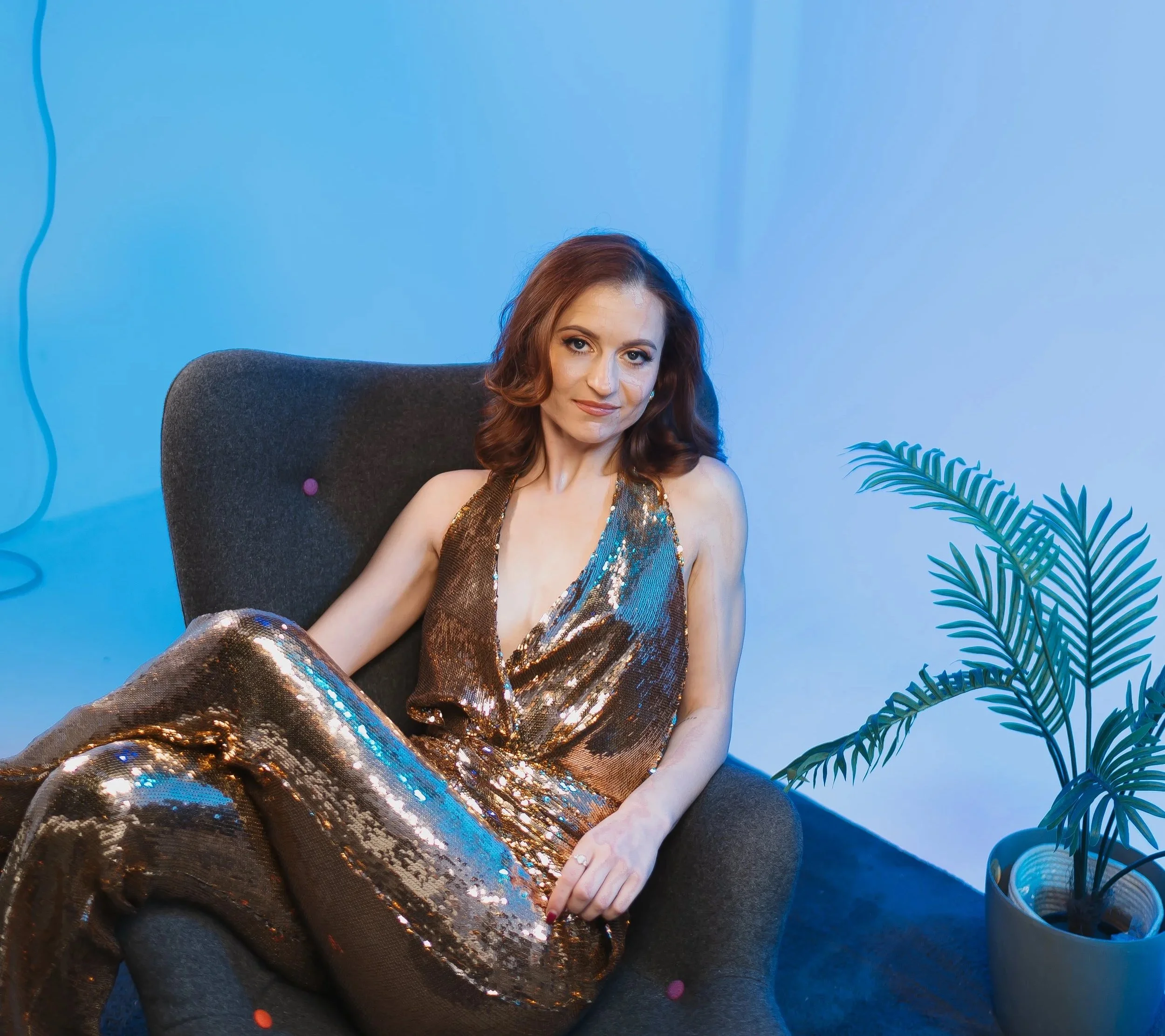 A woman with red hair sitting on a black chair in front of a blue wall, wearing a shiny, gold-colored dress with sequins, next to a green plant.