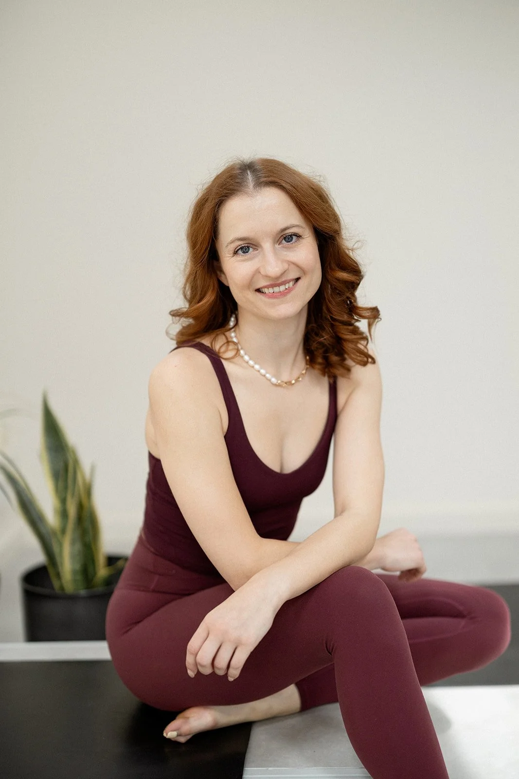 meg mindstrong yoga teacher with red hair wearing a maroon tank top and maroon leggings, sitting cross-legged on a mat indoors with a plant in the background.
