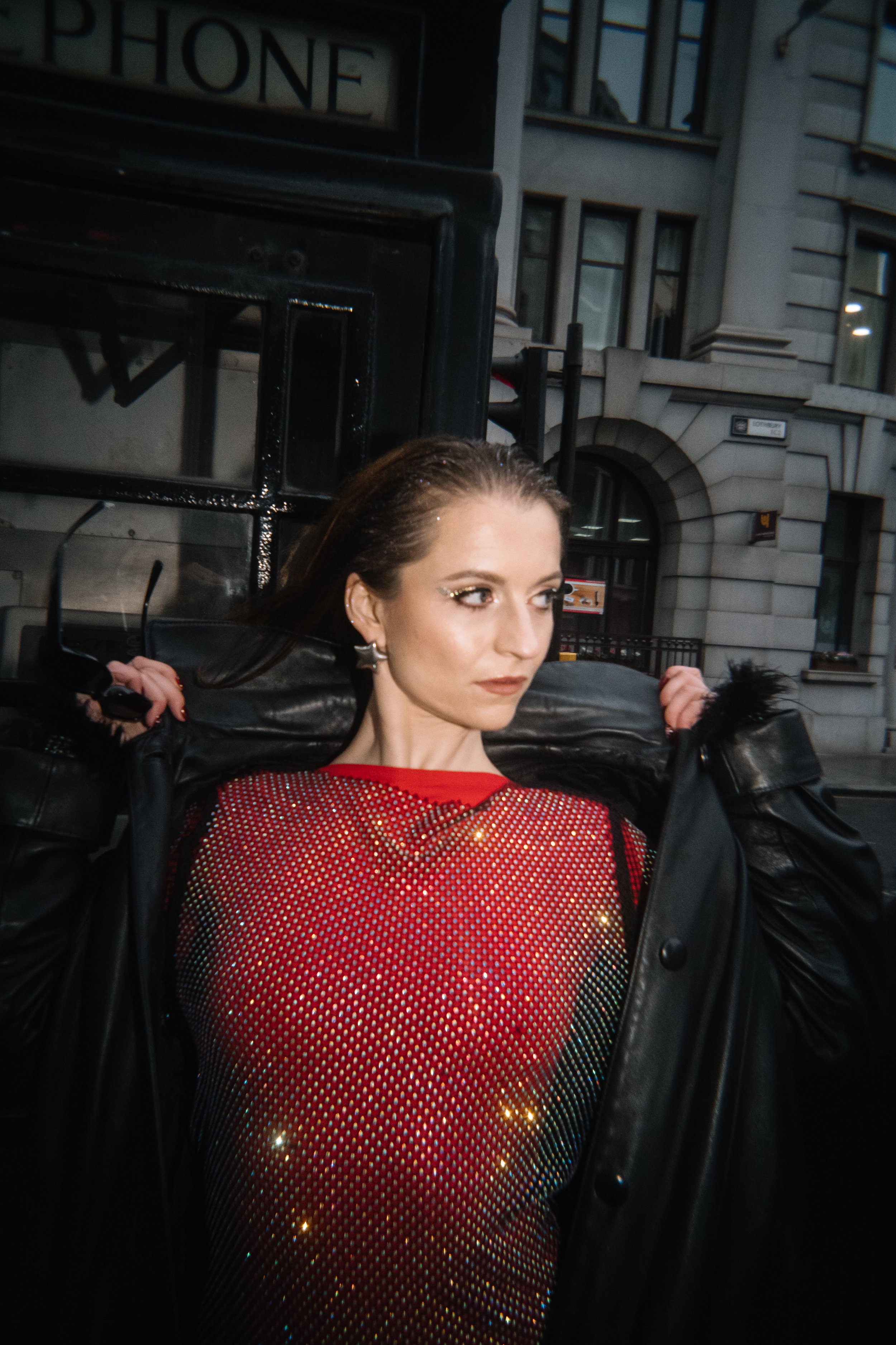 meg mindstrong with dark hair and makeup posing on the street at night, wearing a red sequin dress and a black leather jacket, with city buildings and a phone booth in the background.