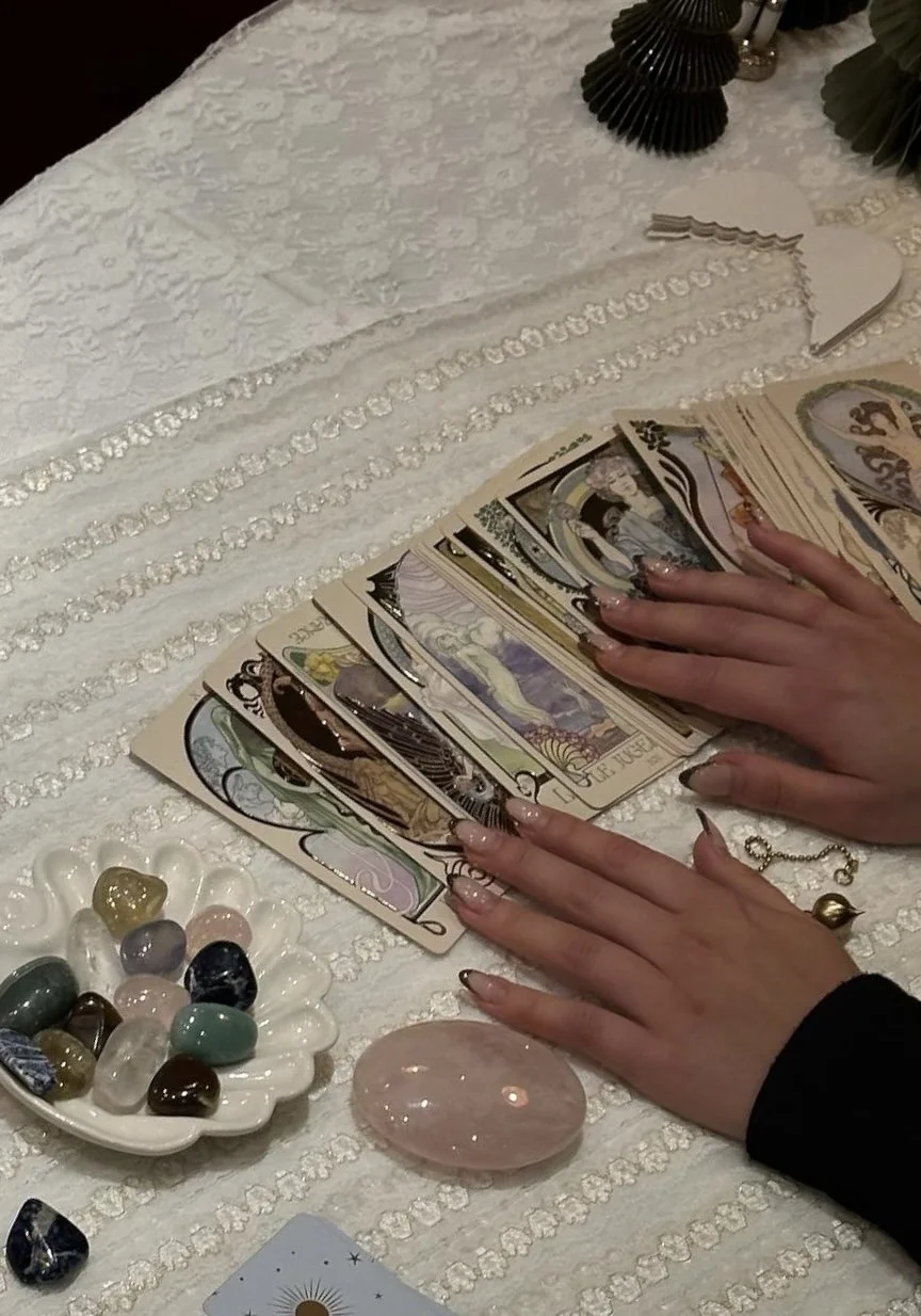 A table covered with a white textured cloth, featuring tarot cards laid out, a plate of polished stones, a large rose quartz crystal, a necklace, and decorative items including black fans and green foliage.