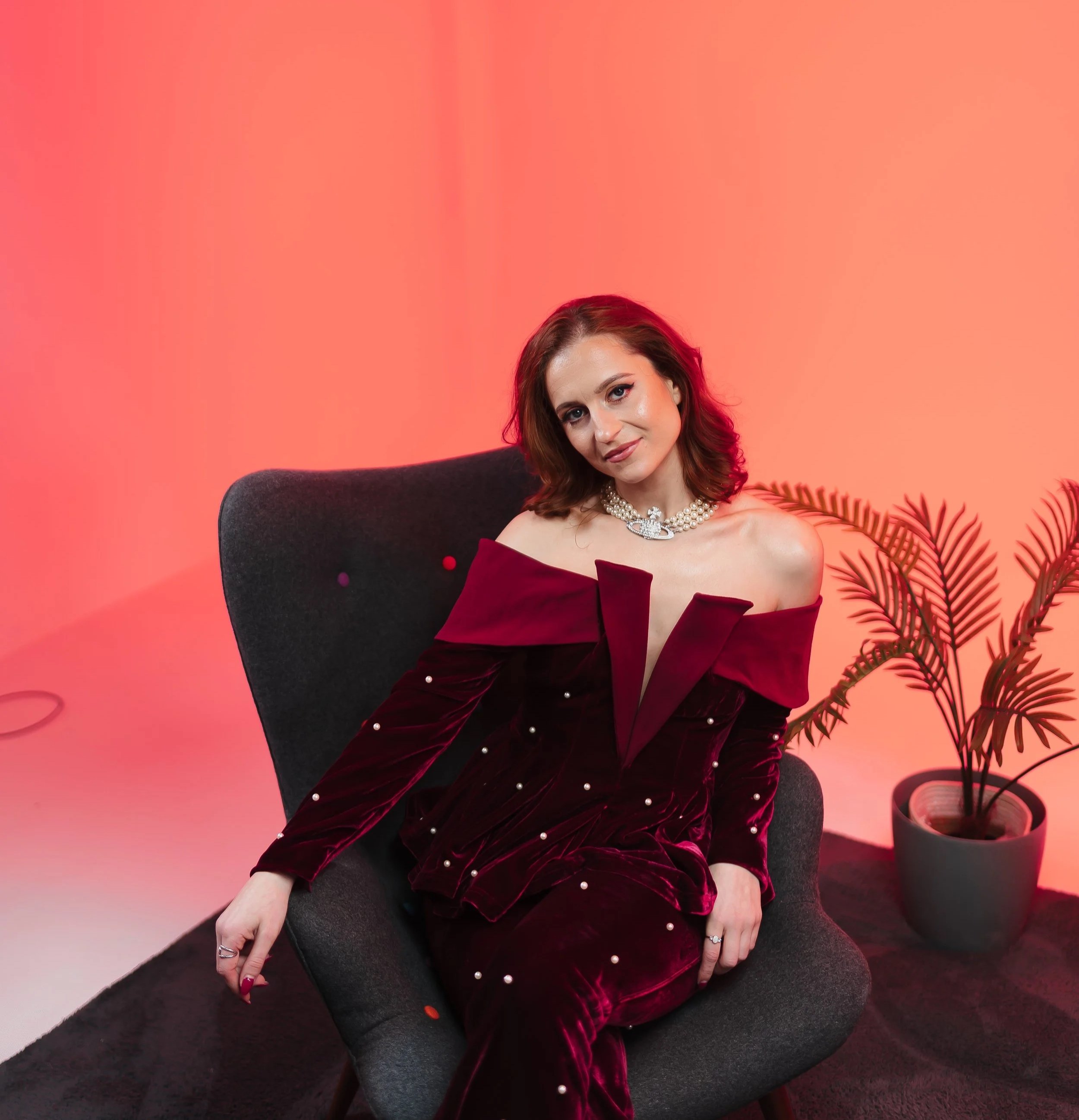 meg mindstrong manifestation coach with shoulder-length brown hair, wearing a dark red velvet off-shoulder dress with pearl embellishments, sitting on a black chair with a red and orange gradient background, and a large green plant to her right.