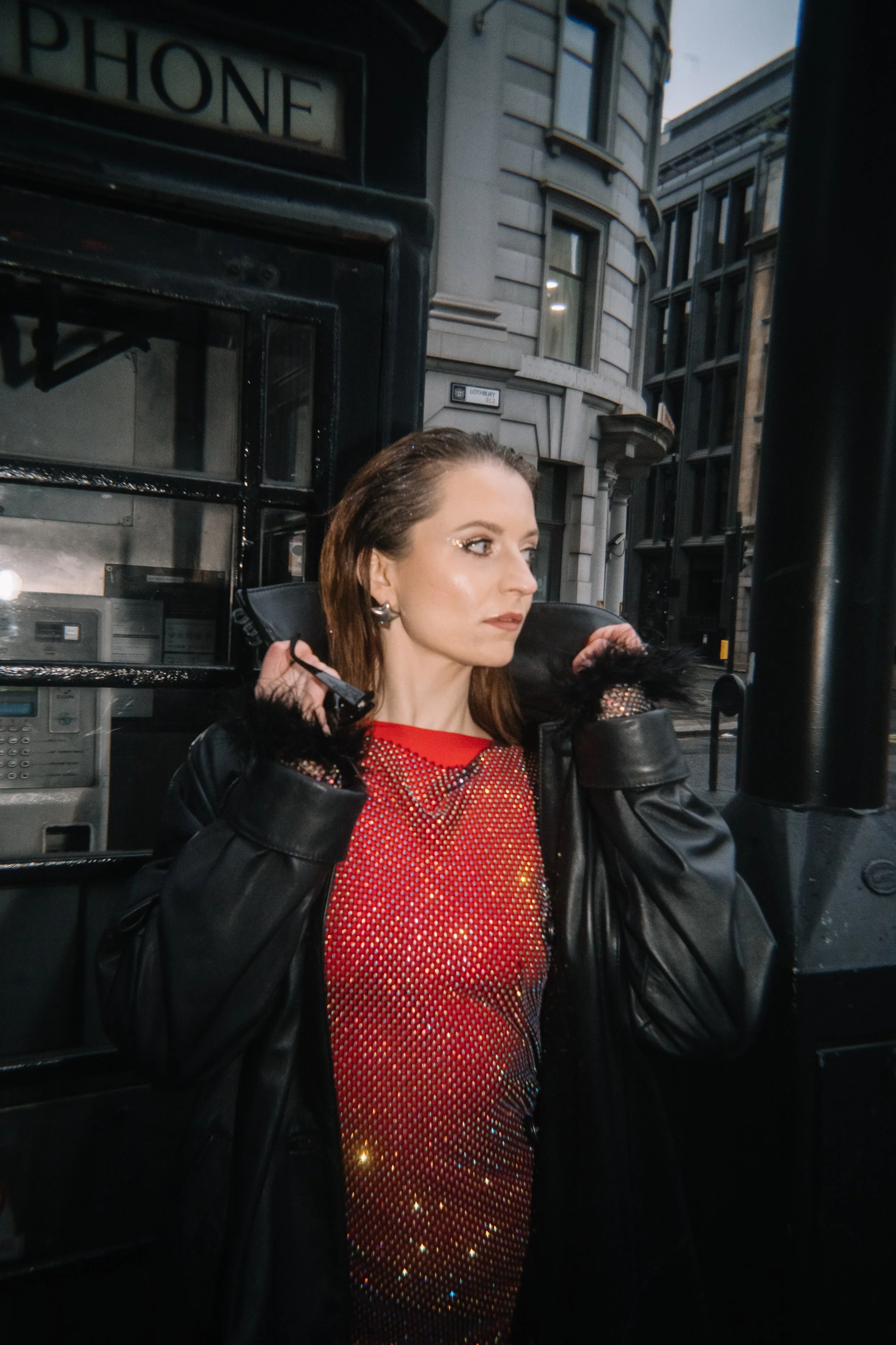 A woman with brown hair and earrings, wearing a black jacket with a fur collar and a red sequined dress, stands near a black phone booth on a city street with old buildings in the background.