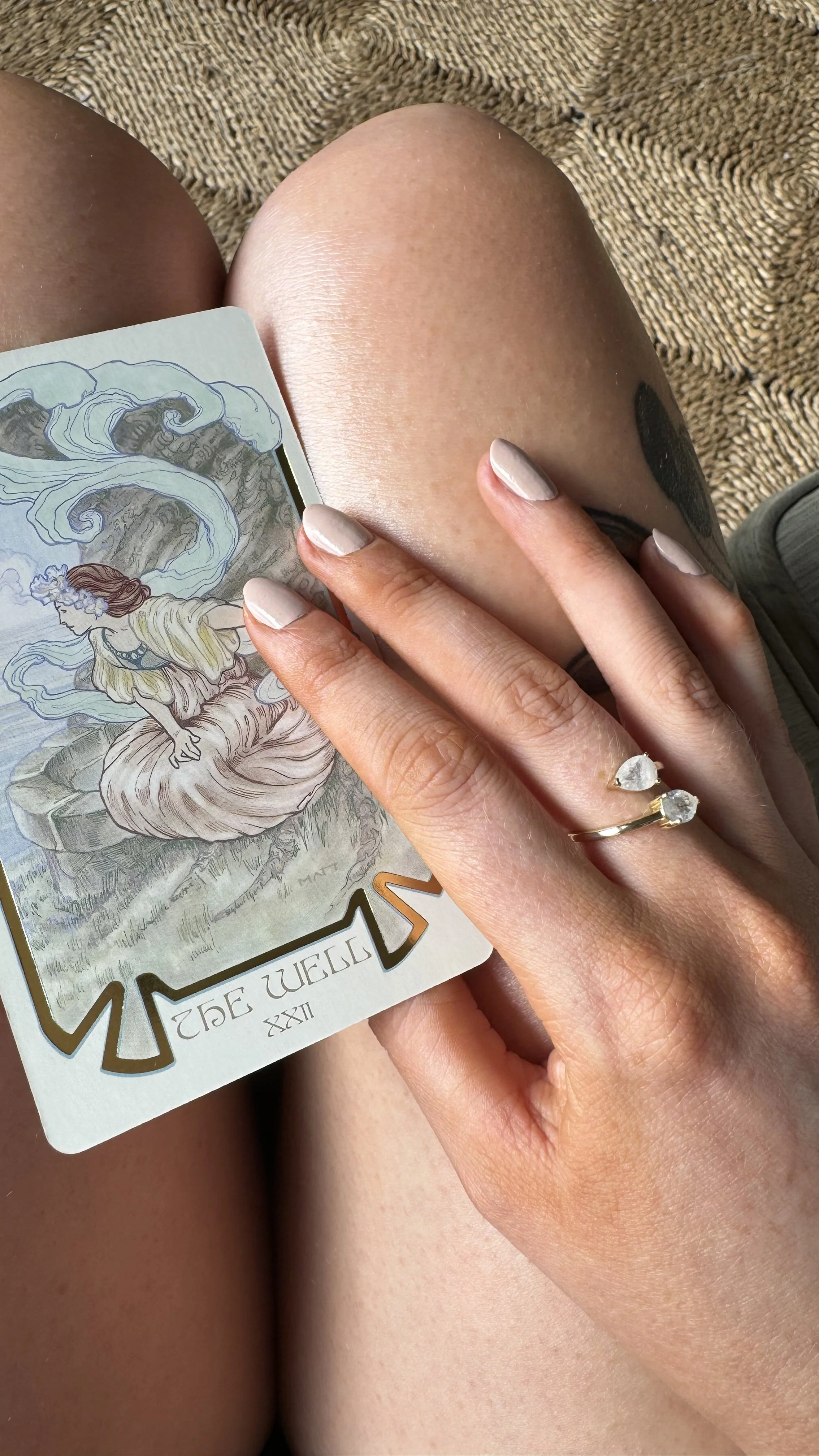 Close-up of a person's legs with a tarot card, The World, held against their thigh. The person has light-colored nail polish, a ring with two clear stones, and a tattoo partially visible on their arm. They are seated on a textured brown woven rug.
