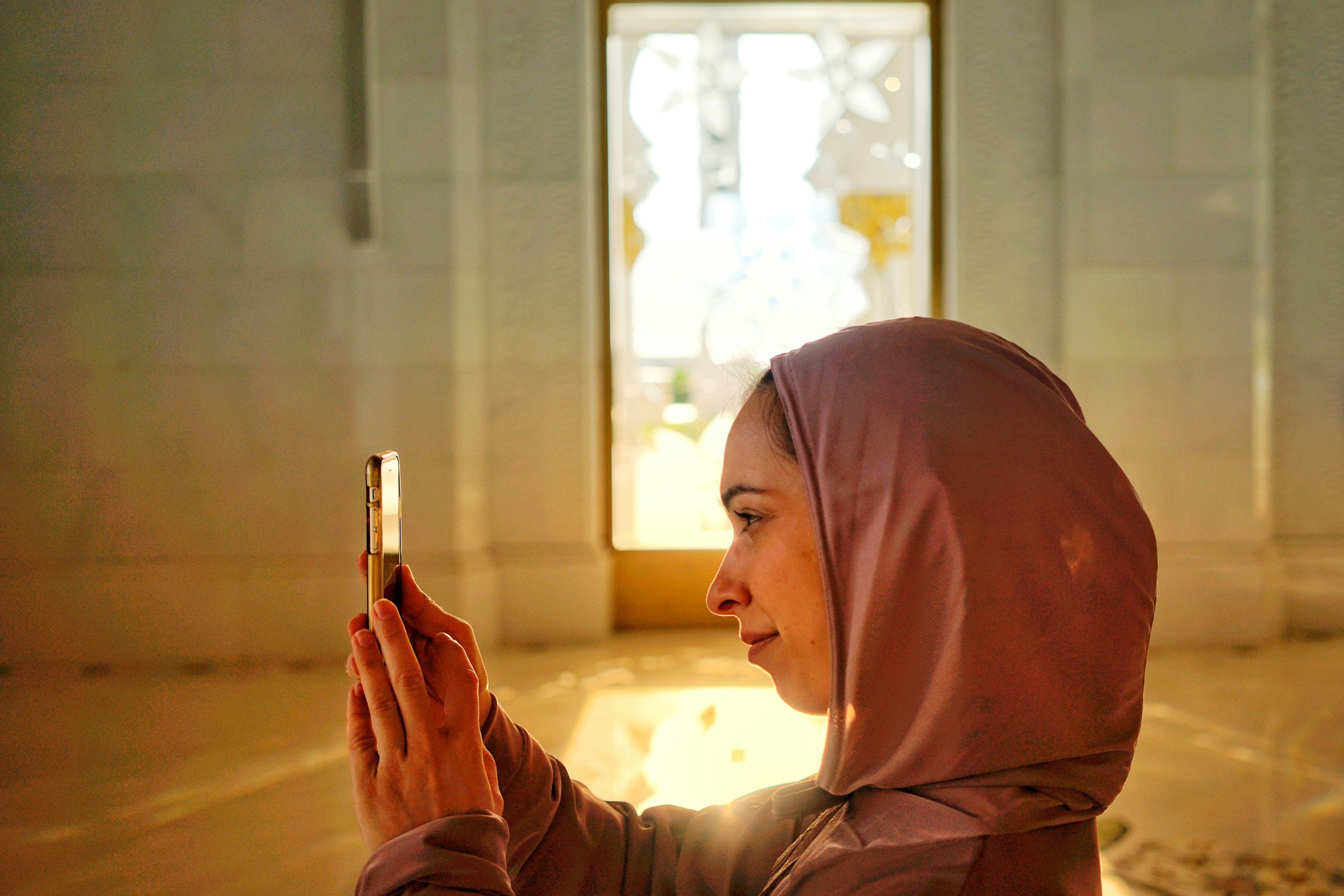 A woman wearing a hijab and smiling while looking at her phone screen.
