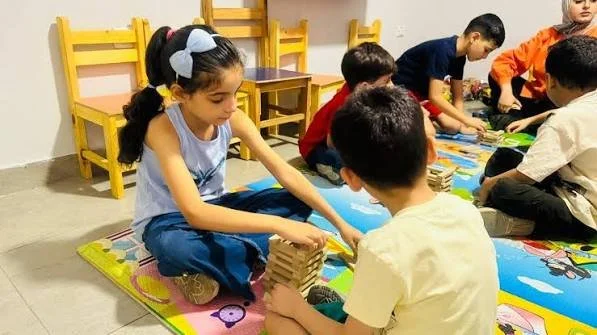 Children sitting on the colorful floor mat and playing with wooden blocks indoors, with yellow chairs in the background.