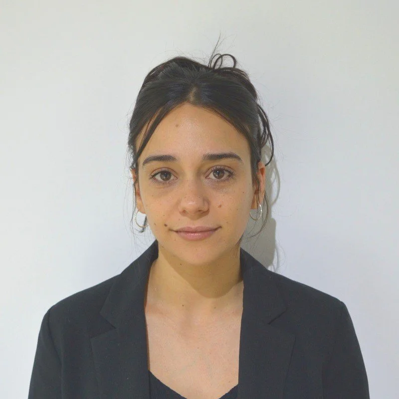 Young woman with dark hair tied back, wearing a black blazer, standing against a plain white wall.
