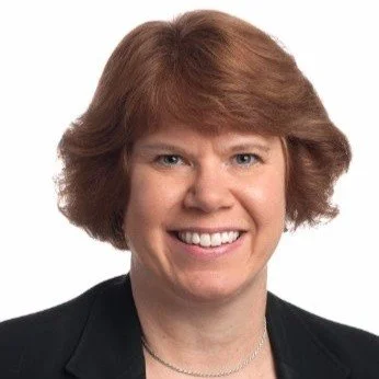 Portrait of a woman with short, curly red hair, smiling, wearing a black blazer and a necklace.